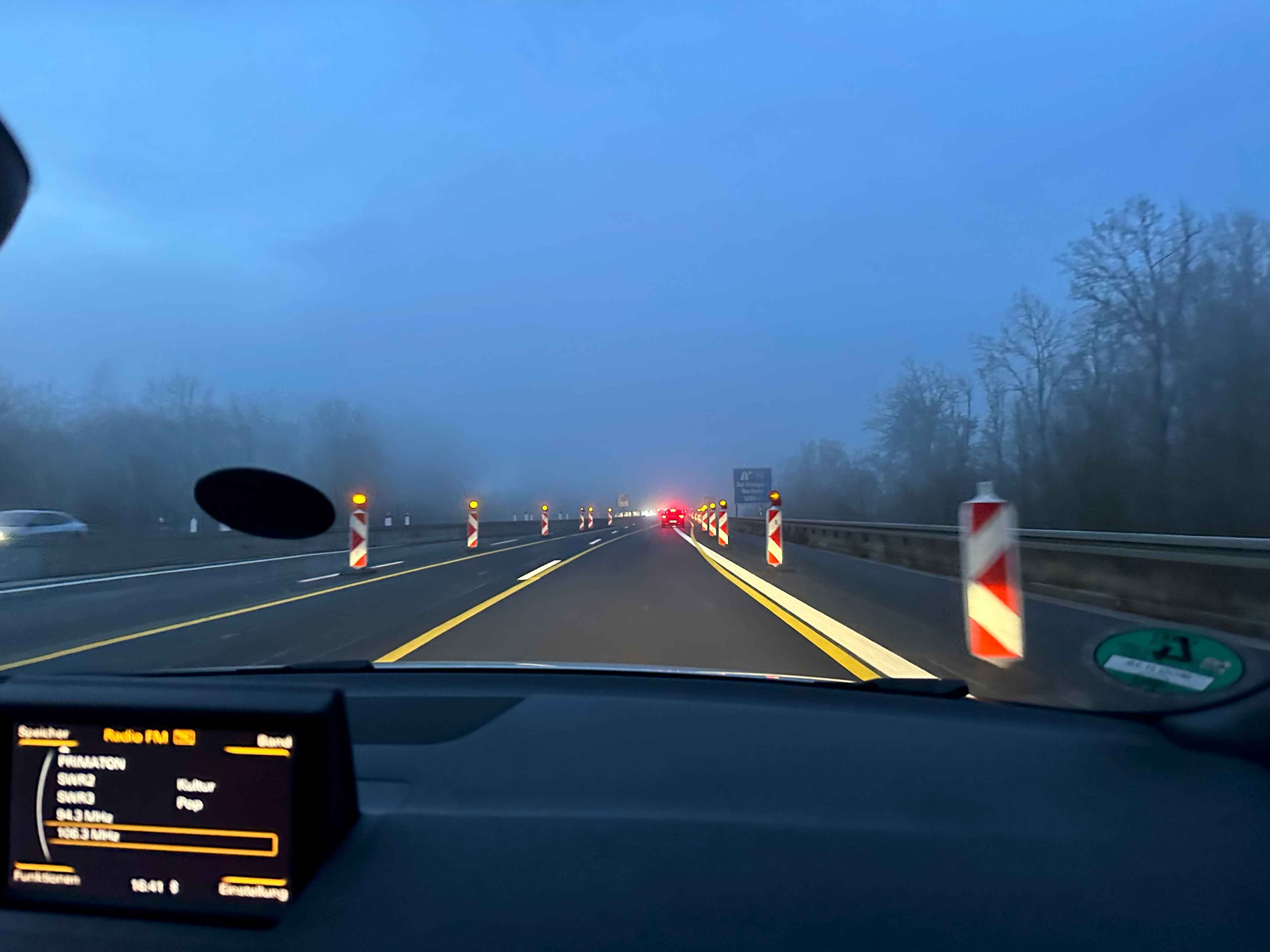 Blick aus einem Auto auf eine neblige Straße bei Nacht mit Baustellenabsperrungen und gelben Fahrbahnmarkierungen.