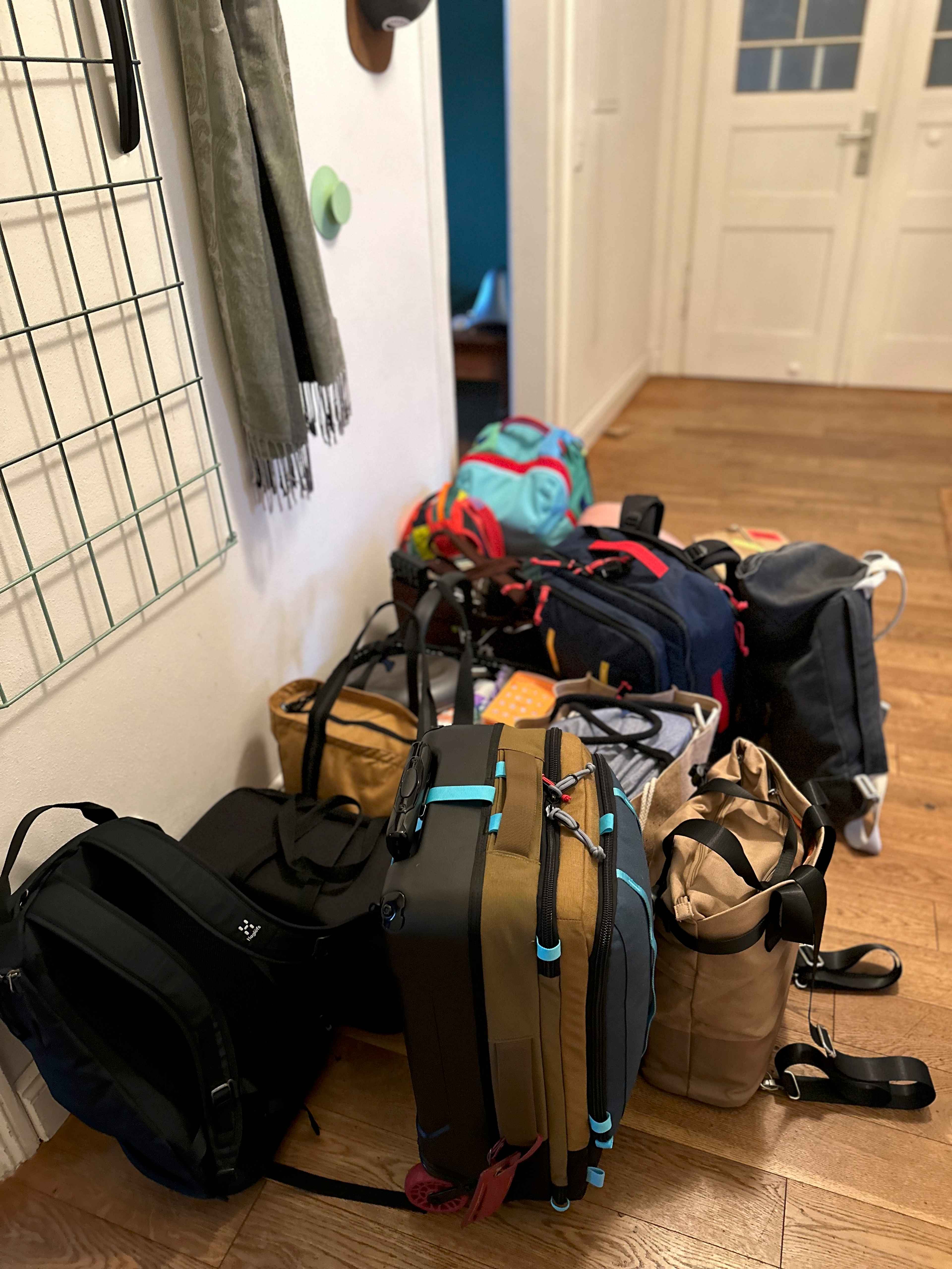 Viele bunte Taschen und Koffer stehen neben einer weißen Wand auf hellbraunem Holzboden im Eingangsbereich einer Wohnung.