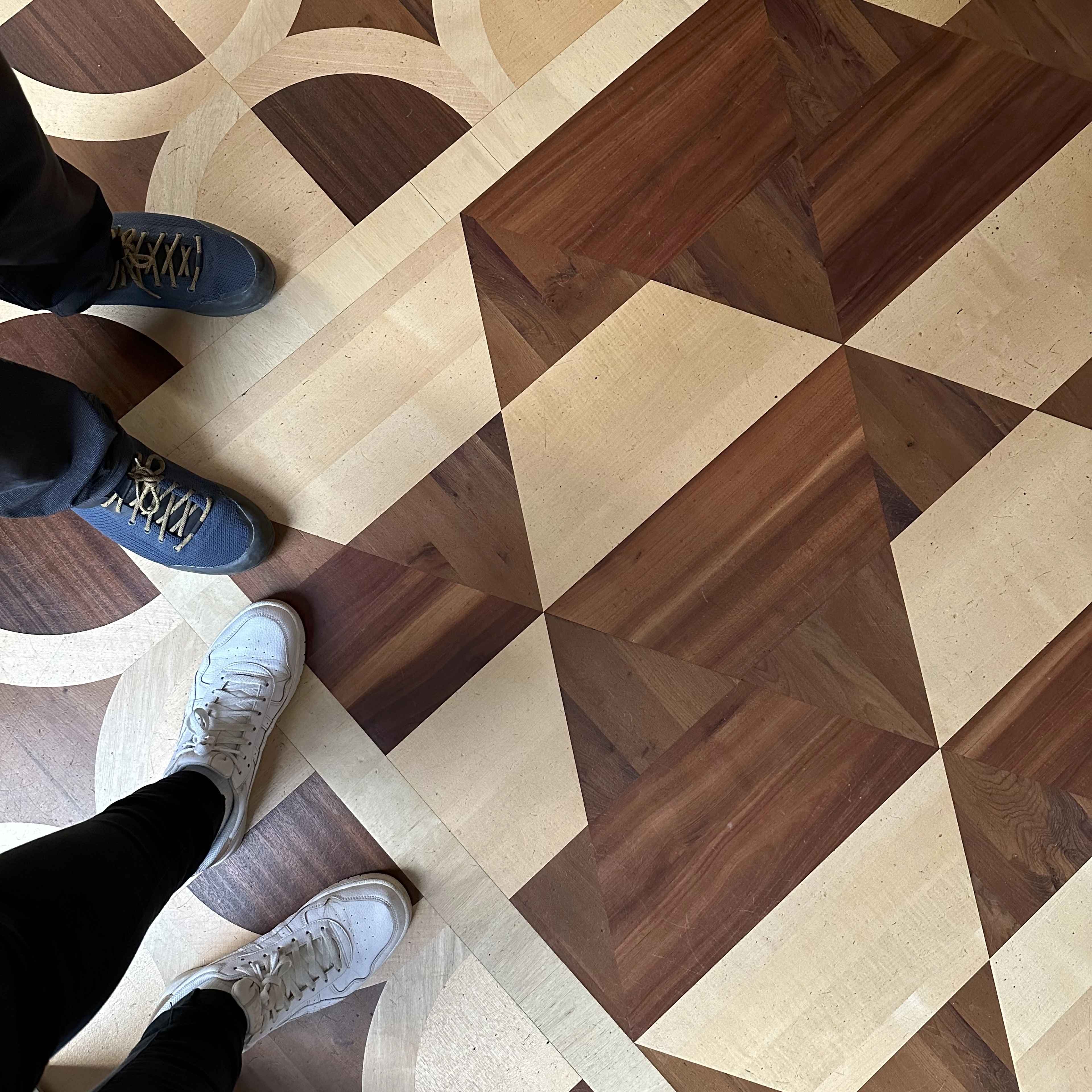 Nur Beine am oberen Rand sichtbar: Zwei Personen stehen auf einem Holzboden mit geometrischen Mustern.