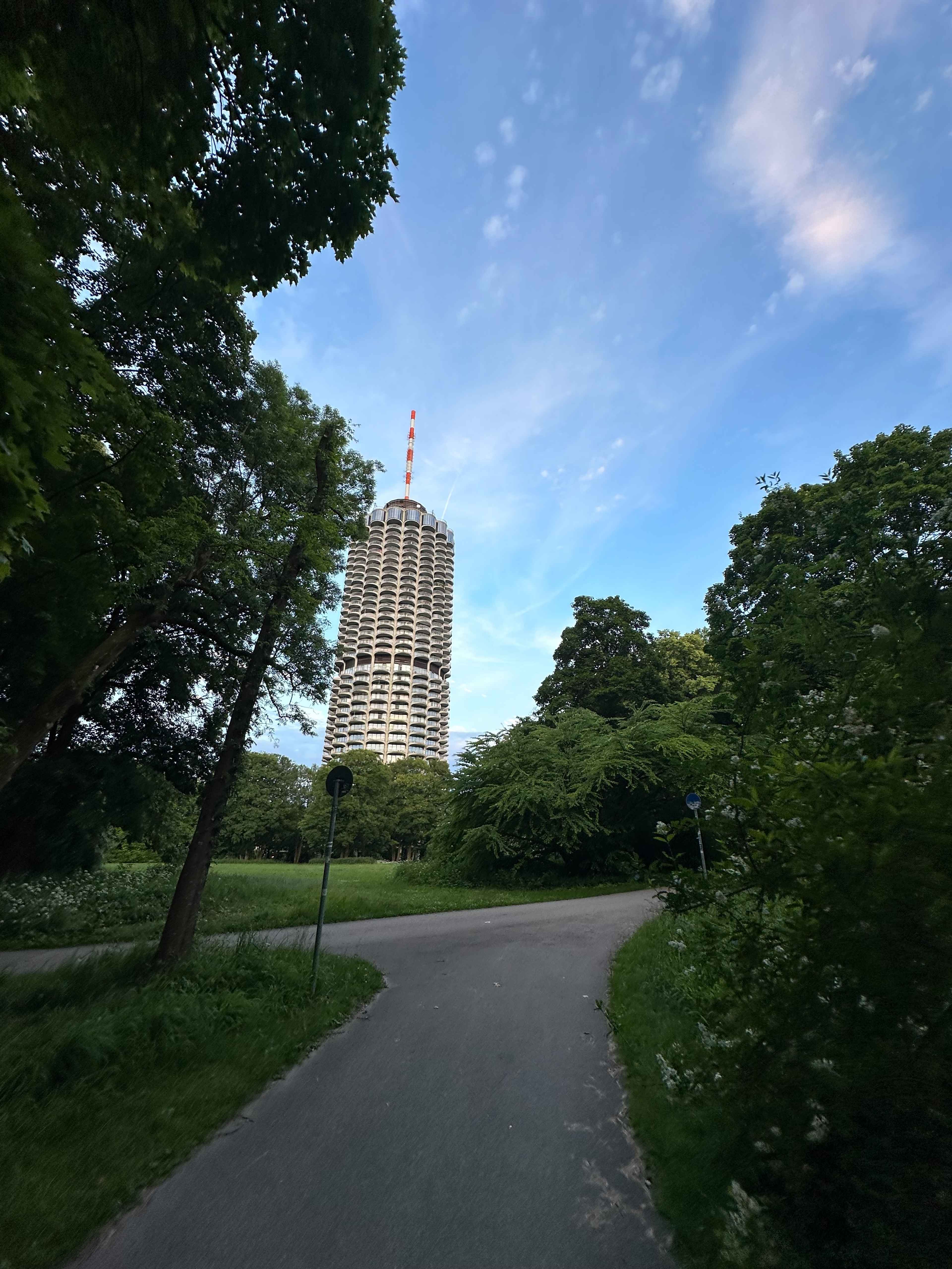 Blick aus dem Park auf den Hotelturm im Hintergrund. Vorne ein sich verzweigender Gehweg und grüne Bäume an den Seiten.