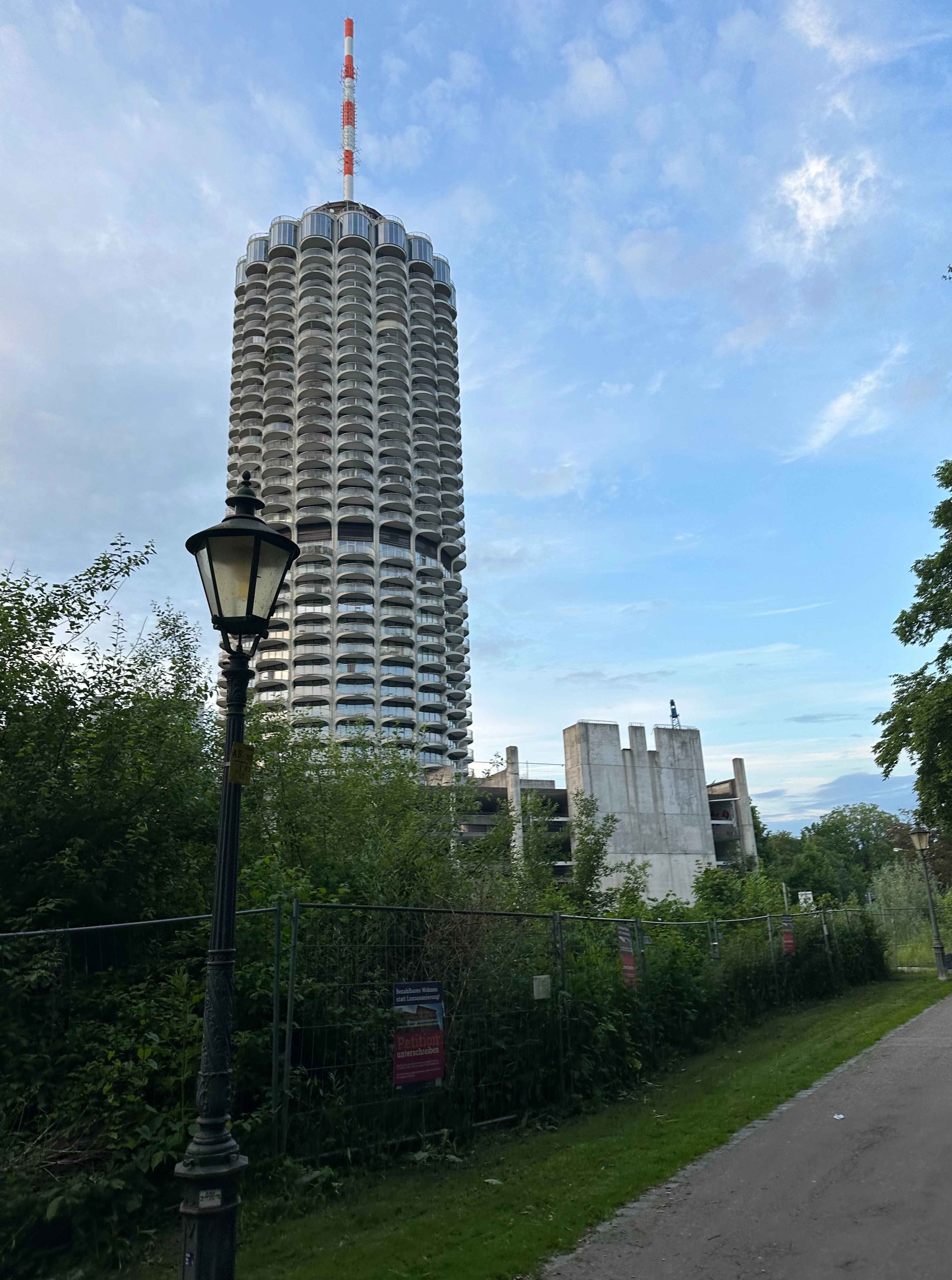 Blick auf den Augsburger Hotelturm. Im Vordergrund eine historische Straßenlaterne, grüne Büsche und ein Bauzaun.