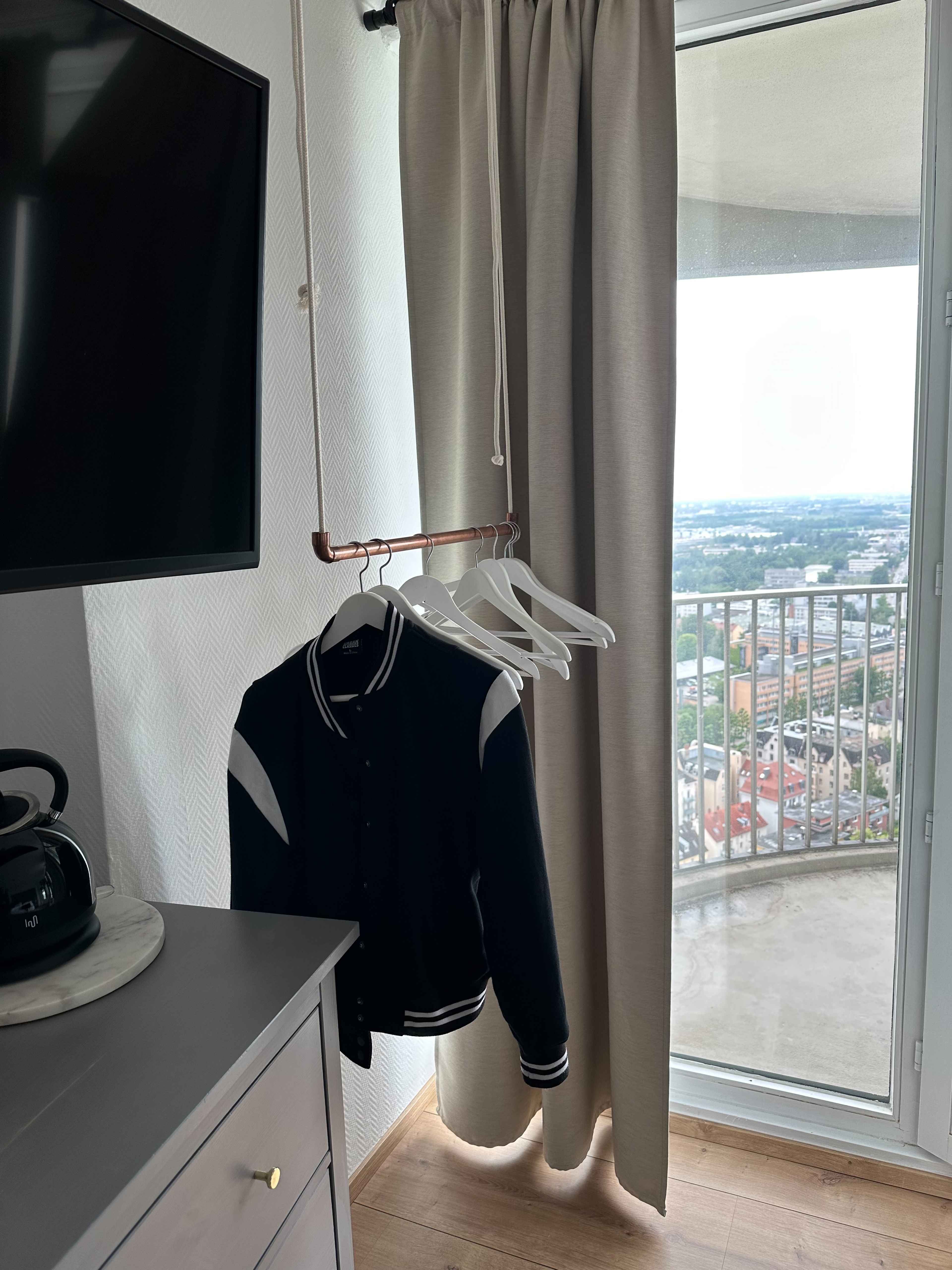 Zimmer im Hotelturm mit Ausblick, Fernseher und Jacke an einem Kleiderbügel vor einem Fenster.