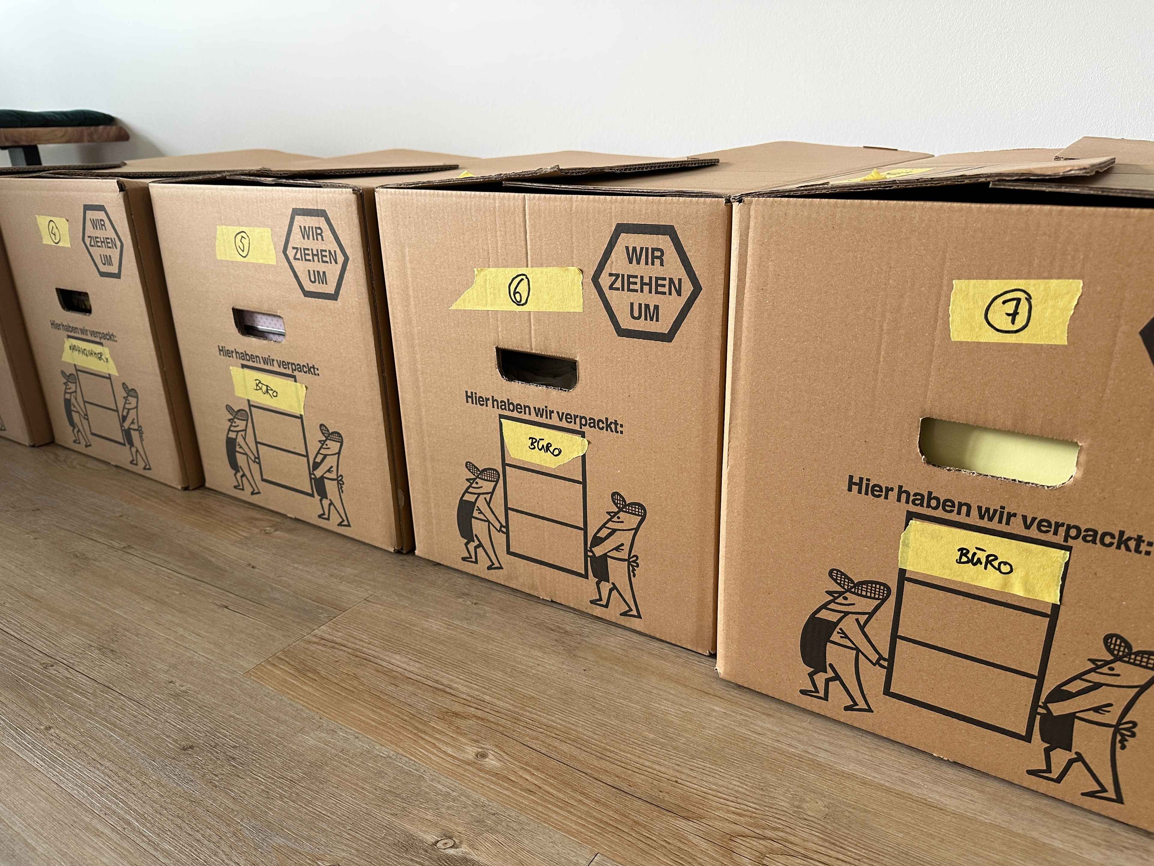 Nahaufnahme einer Reihe Umzugskartons mit gelbem Tape und Beschriftung mit Zahl und Raumname.
