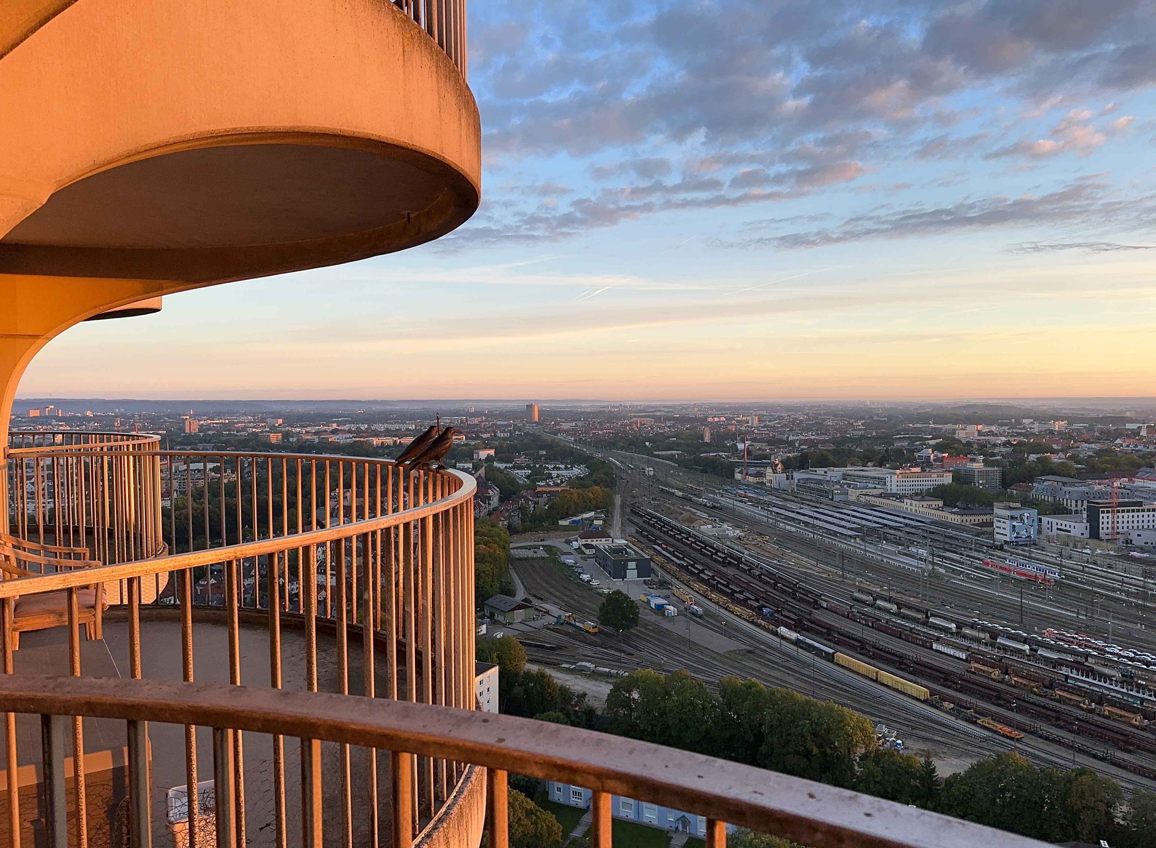 Aussicht aus dem 31. Stock des Hotelturms im Abendlicht. Links ein runder Balkon mit Raben-Figuren und Bahnhof im Hintergrund