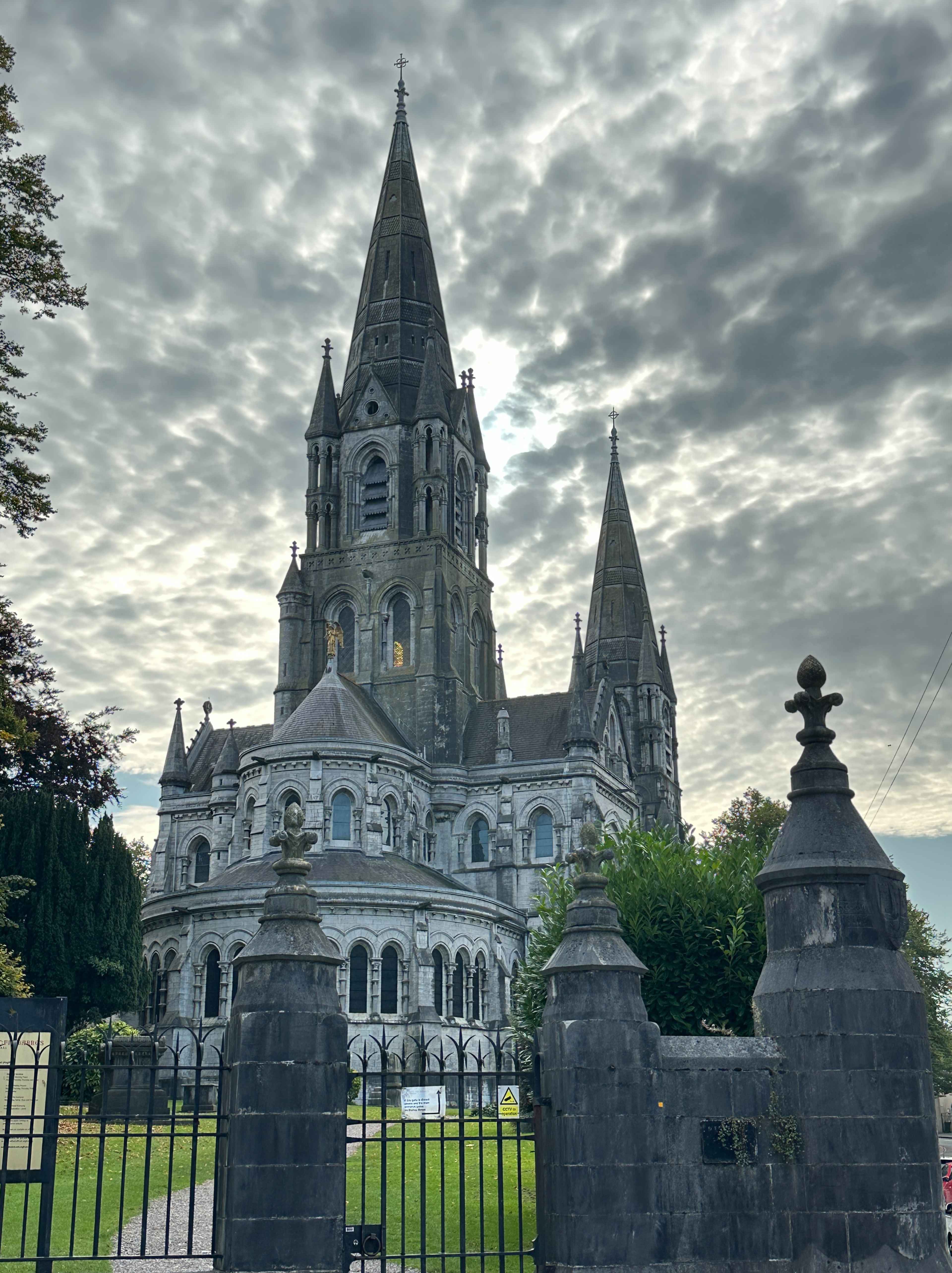 Sicht auf Saint Fin Barre's Cathedral, Cork. Eine graue neogotische Kirche mit spitzen Türmen vor dramatischem Wolkenhimmel.