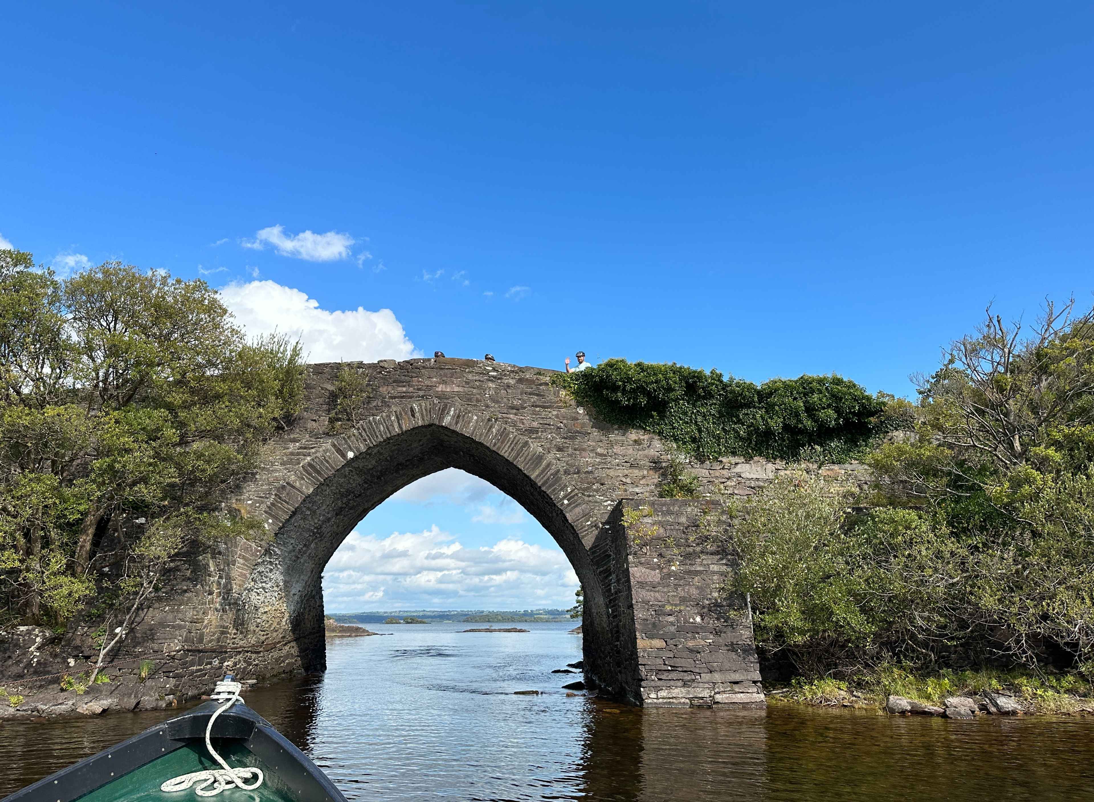 Blick vom Boot aus auf historische Steinbrücke in Dundag Bay mit winkendem Passant unter klarem blauem Himmel.