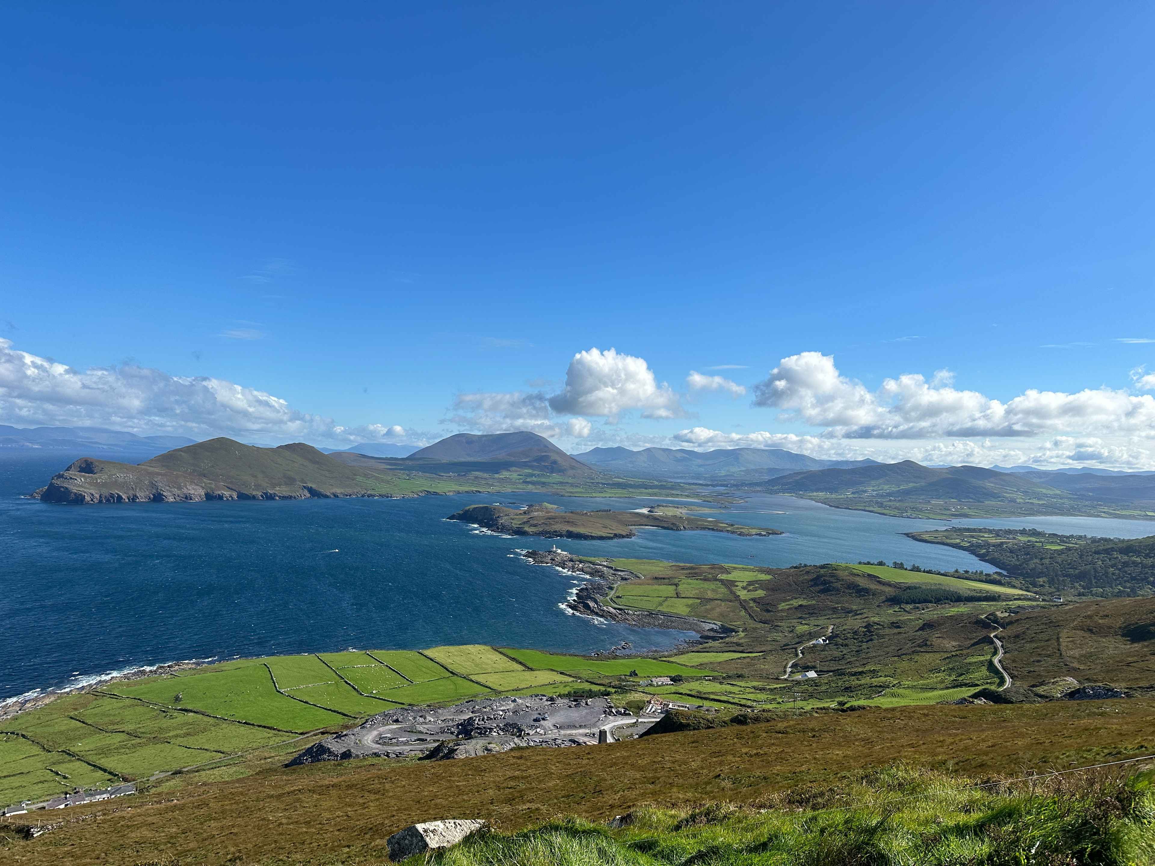 Panorama vom Berg aus auf die Küste von Valentia Island, mit grün-schattierten Feldern, blauem Meer unter blauem Himmel.