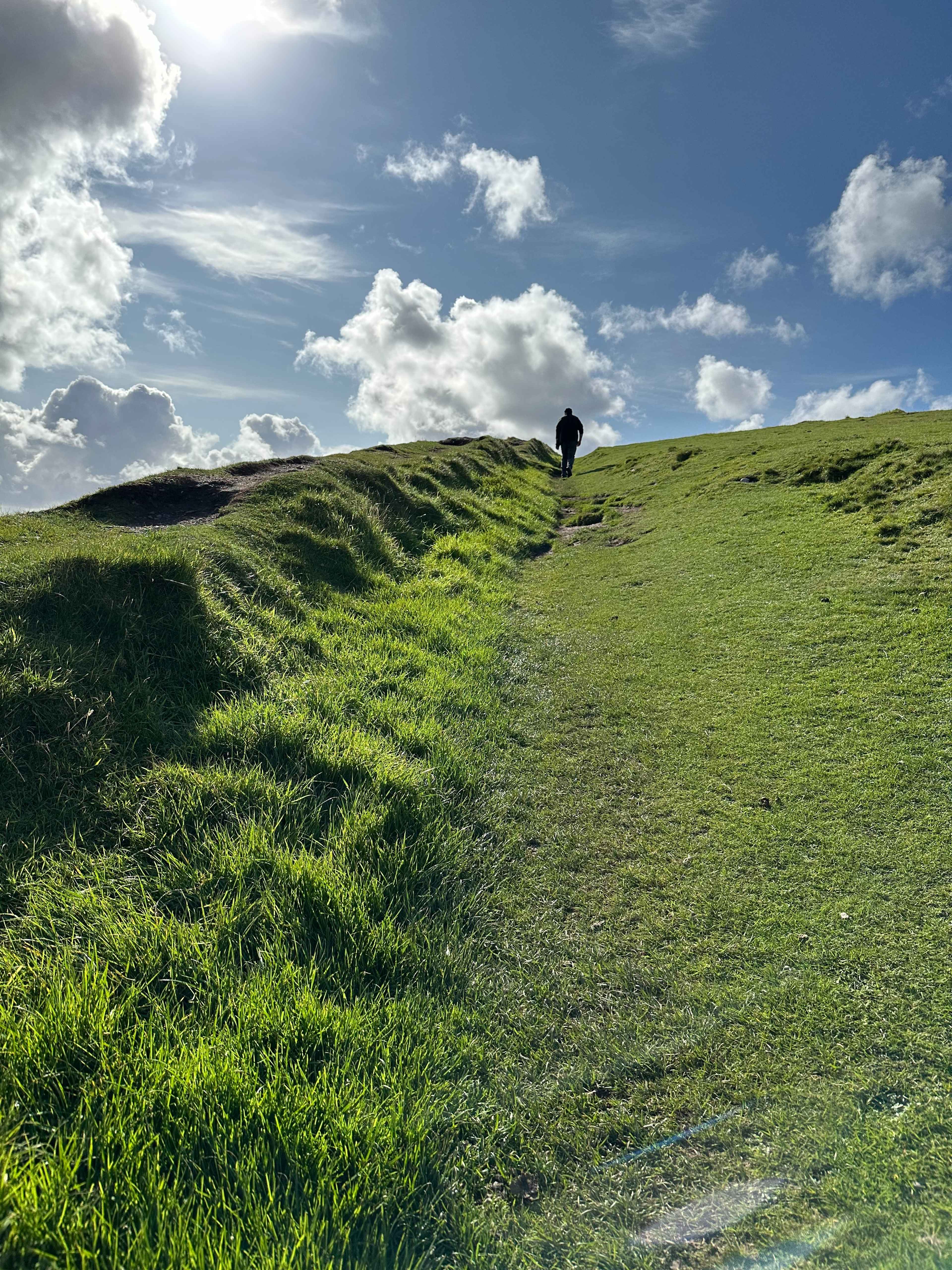 Basti weit hinten geht einen steilen grünen Hügel am Wild Atlantic Way hinauf, umgeben von grünem Gras unter sonnigen Himmel.