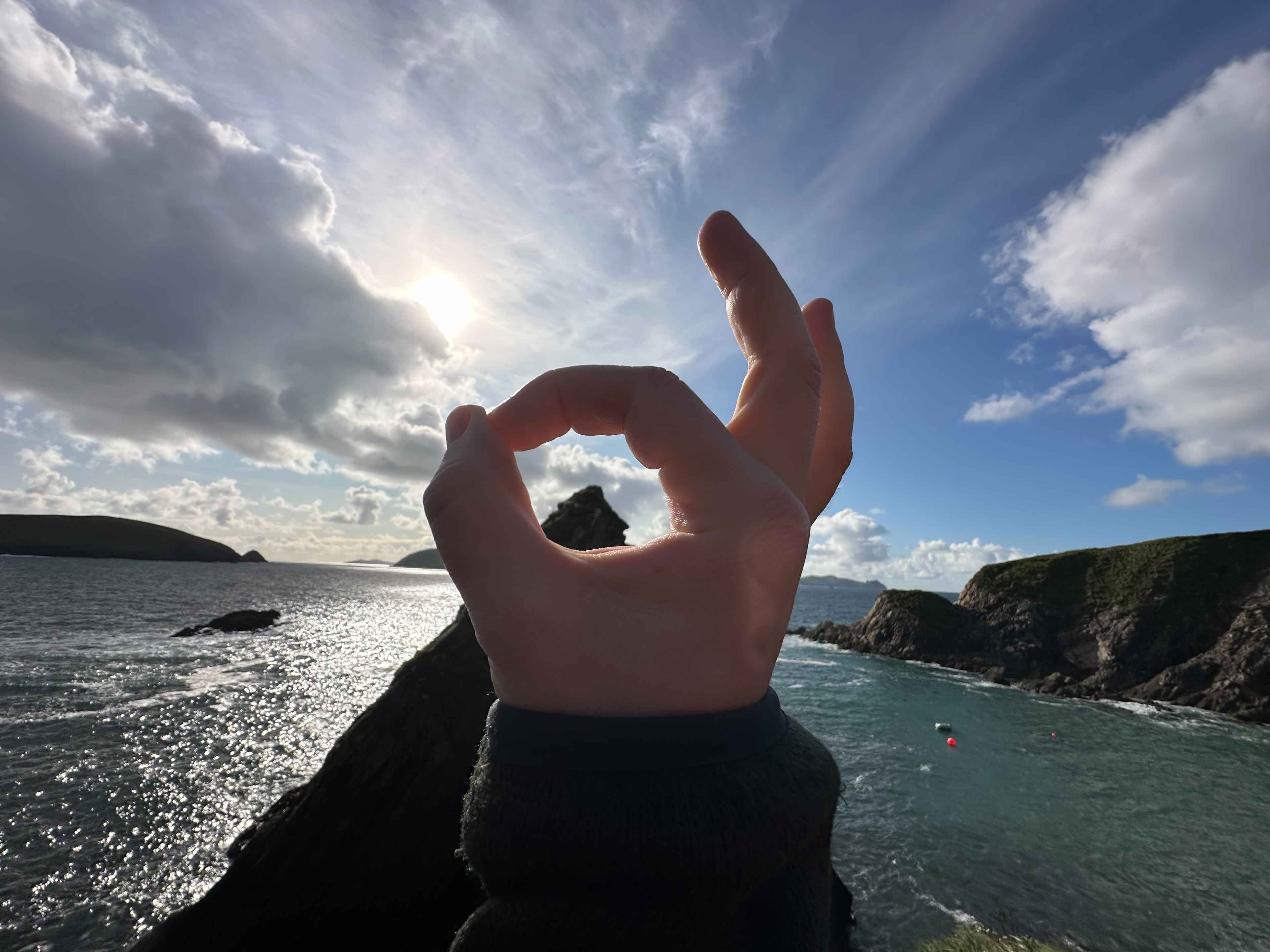 Panorama des Dunquin Pier: Vorne eine Hand in der ‚OK‘-Geste, die die Spitze des von Meer umgebenen Bergs einrahmt.