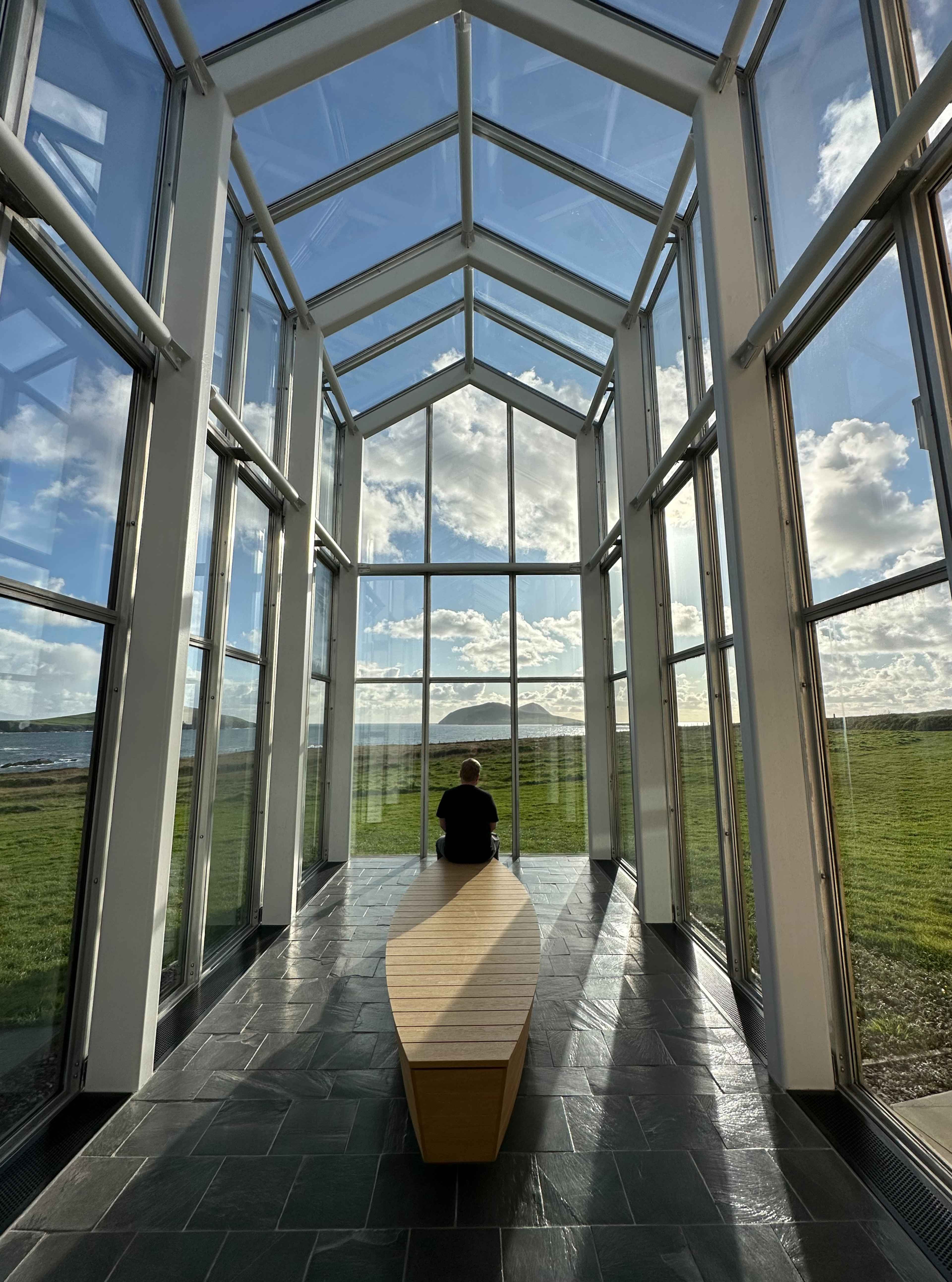 Lichtdurchflutetes Glasgebäude im Blasket Center mit Blick auf die Küste. Basti von hinten auf einer langen Holzbank sitzend.