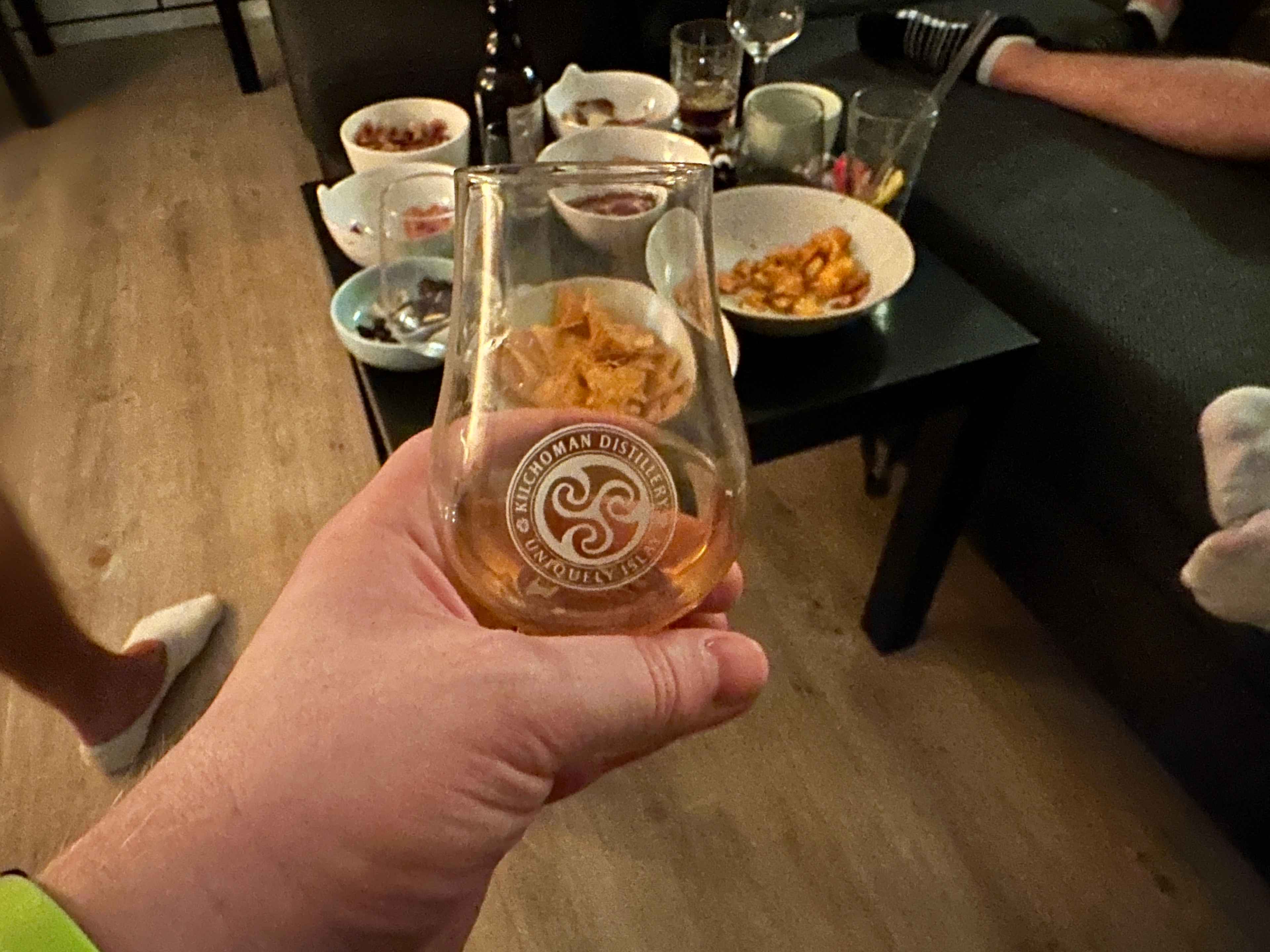 Nahaufnahme einer Hand, die ein befülltes Whiskyglas hält. Im Hintergrund ein Tisch mit weißen Schalen mit Snacks.