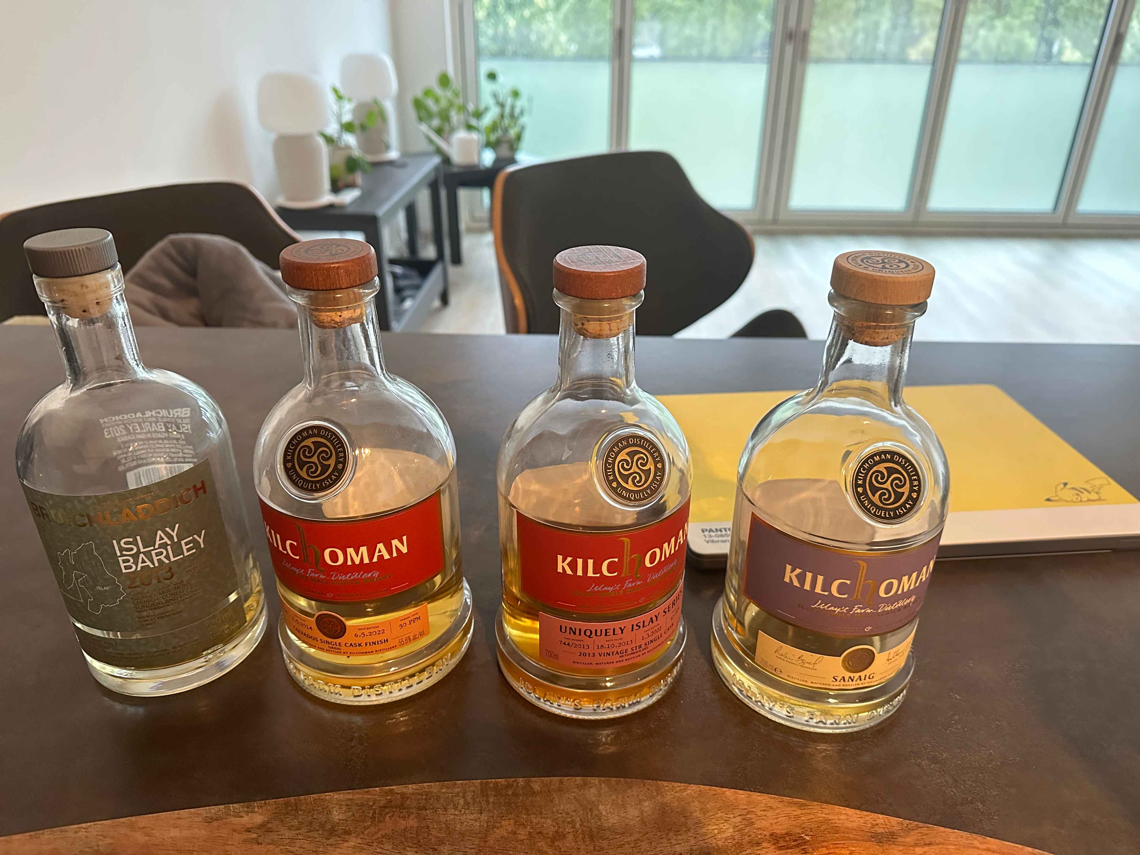 Aufgereihte Kilchoman Whiskyflaschen auf einer dunklen Küchenbank. Dahinter ein gelber Laptop. Mit Fenstern im Hintergrund.