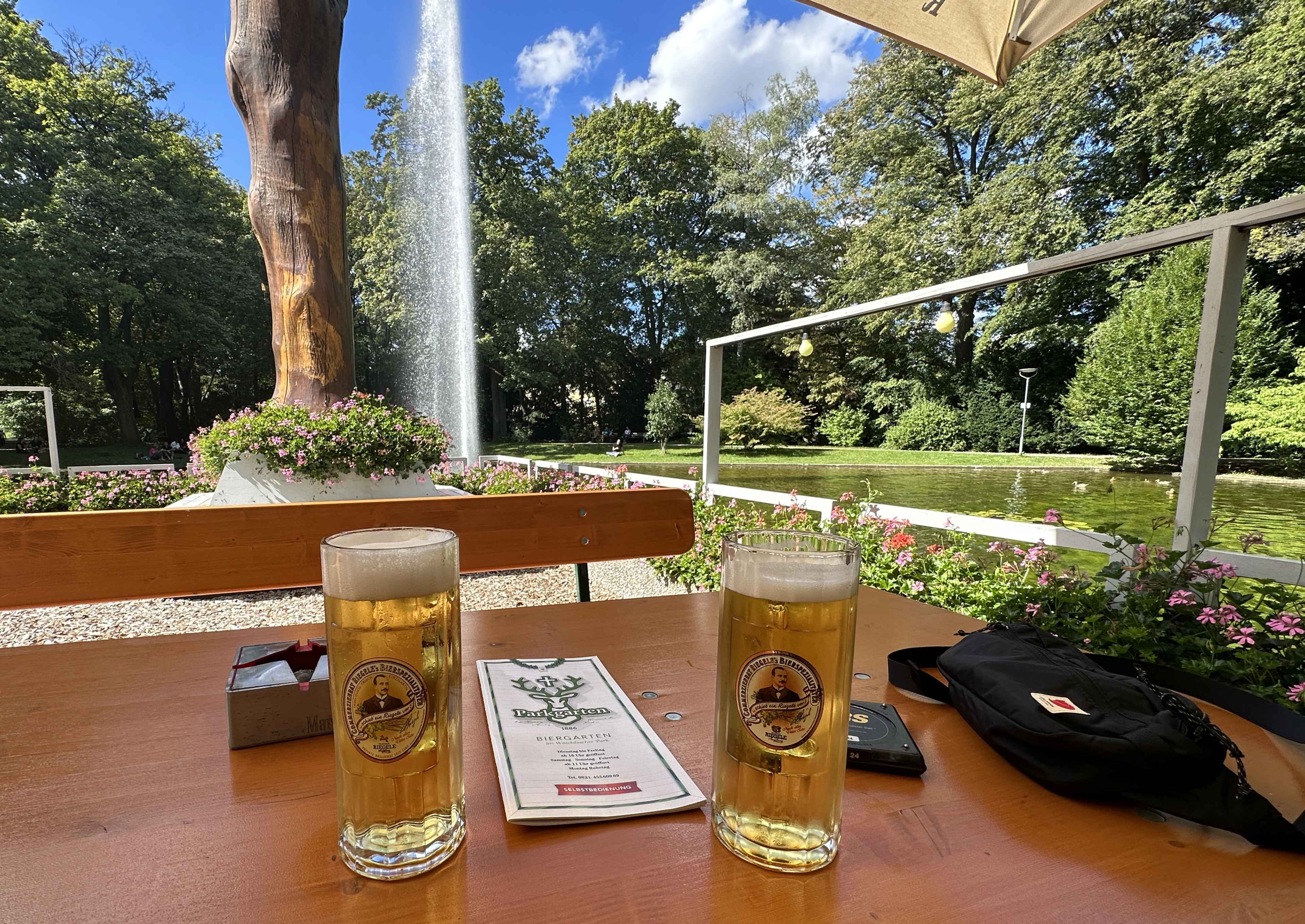 Zwei volle Bierkrüge auf einem Biertisch im Parkgarten Augsburg. Im Hintergrund grüner Park und die Wasserfontäne des Teichs.