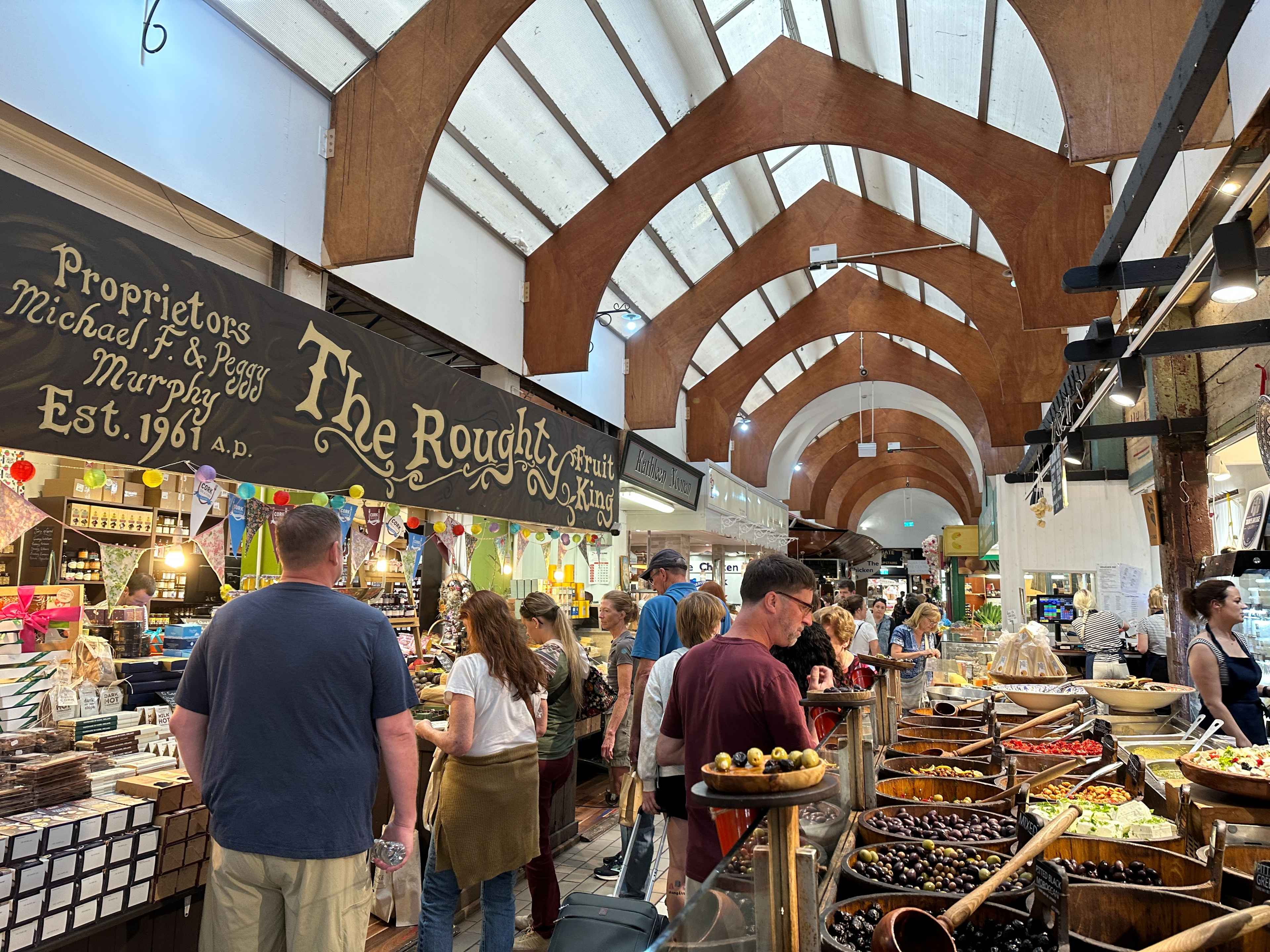 Ein Gang im English Market in Cork mit vielen Leuten. Läden mit ausgestelltem Essen an den Seiten, wie Schüsseln mit Oliven.
