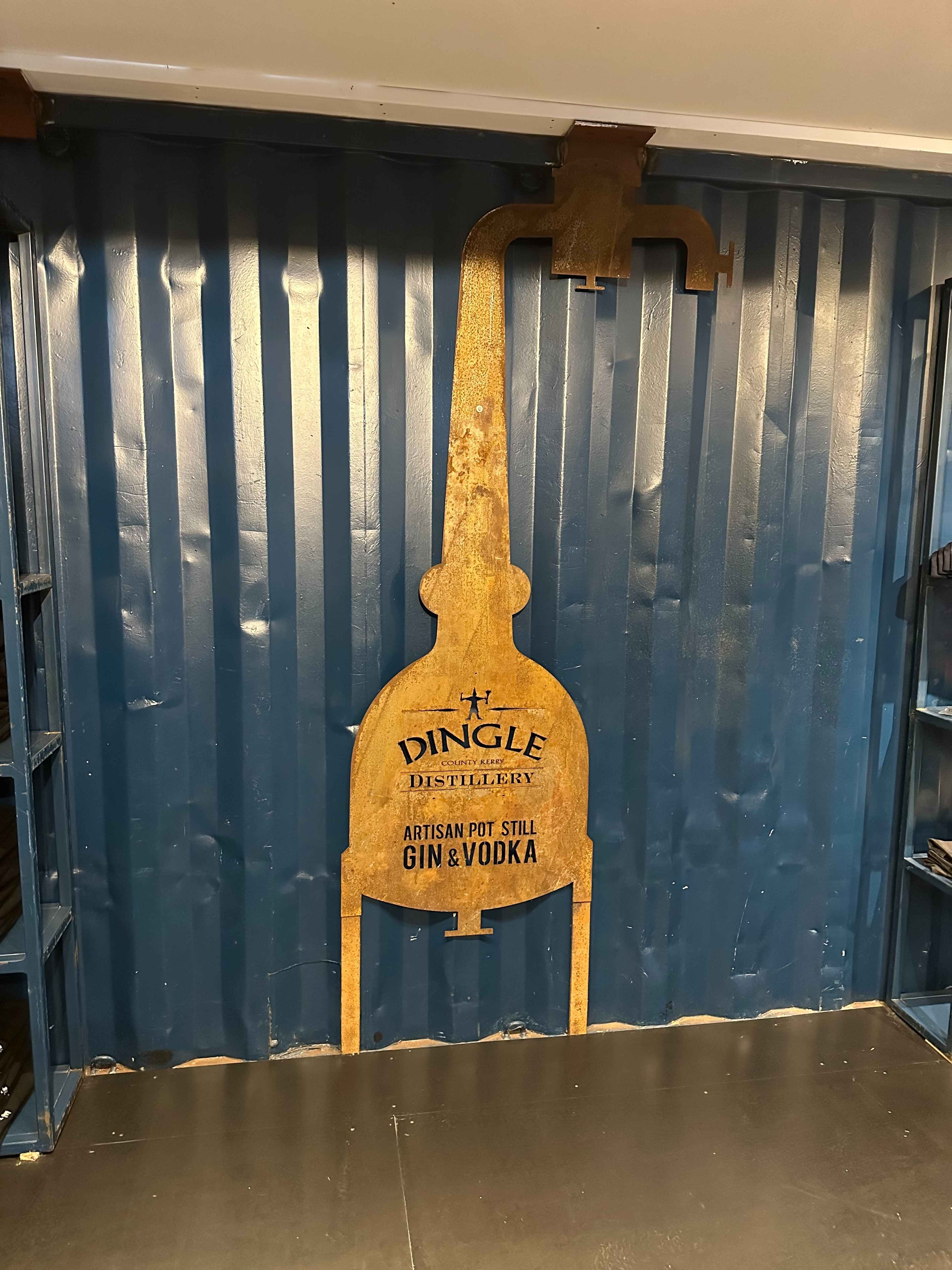 Goldenes Metallschild in Form eines Destillierapparats mit Aufschrift „Dingle Distillery“ auf einer blauen Wellblech-Wand.
