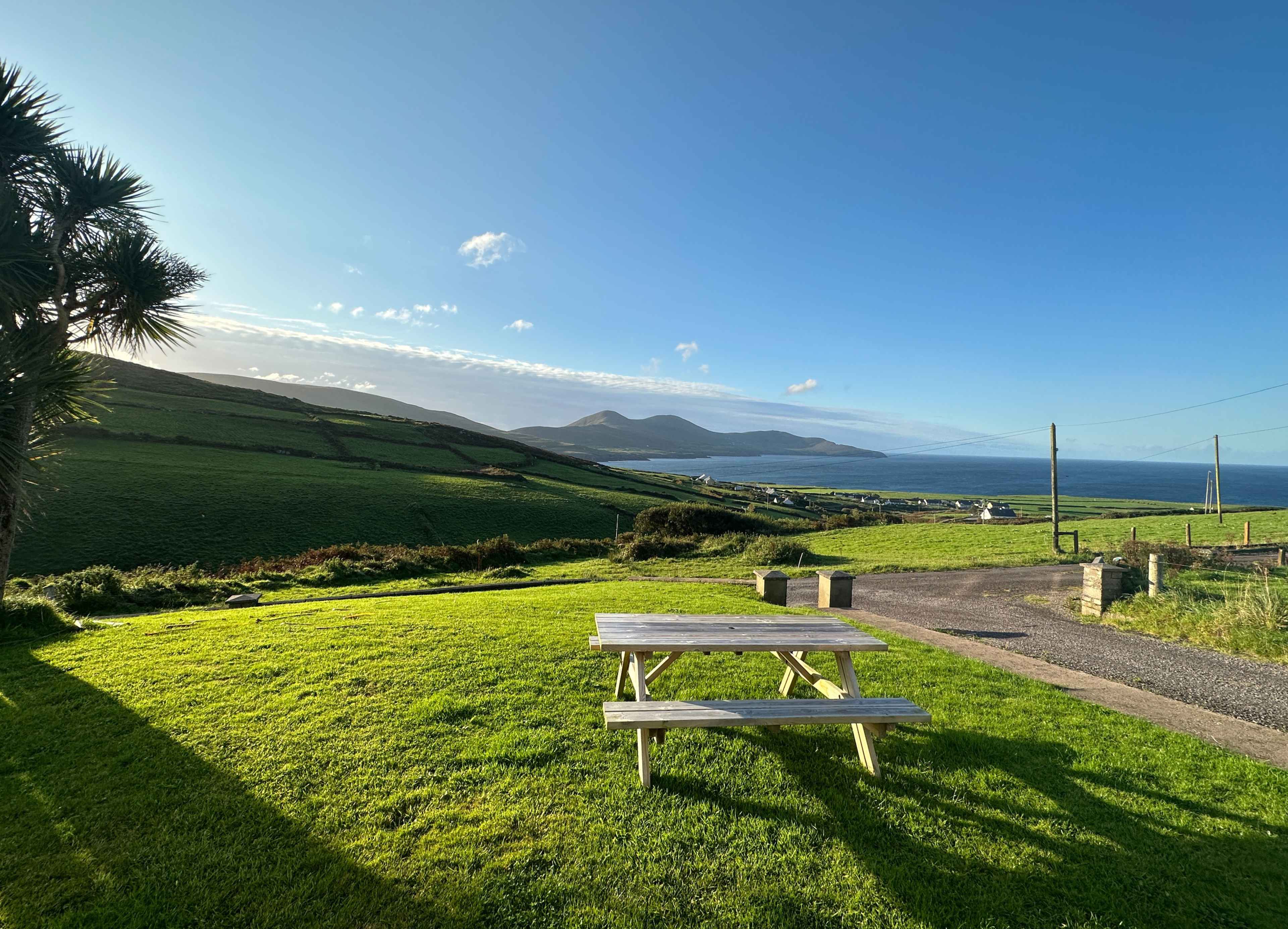 Hügellandschaft mit grünem Gras in The Glen, Kerry. Im Hintergrund rechts das Meer. Vorne: Tisch aus Holz und eine Ausfahrt.