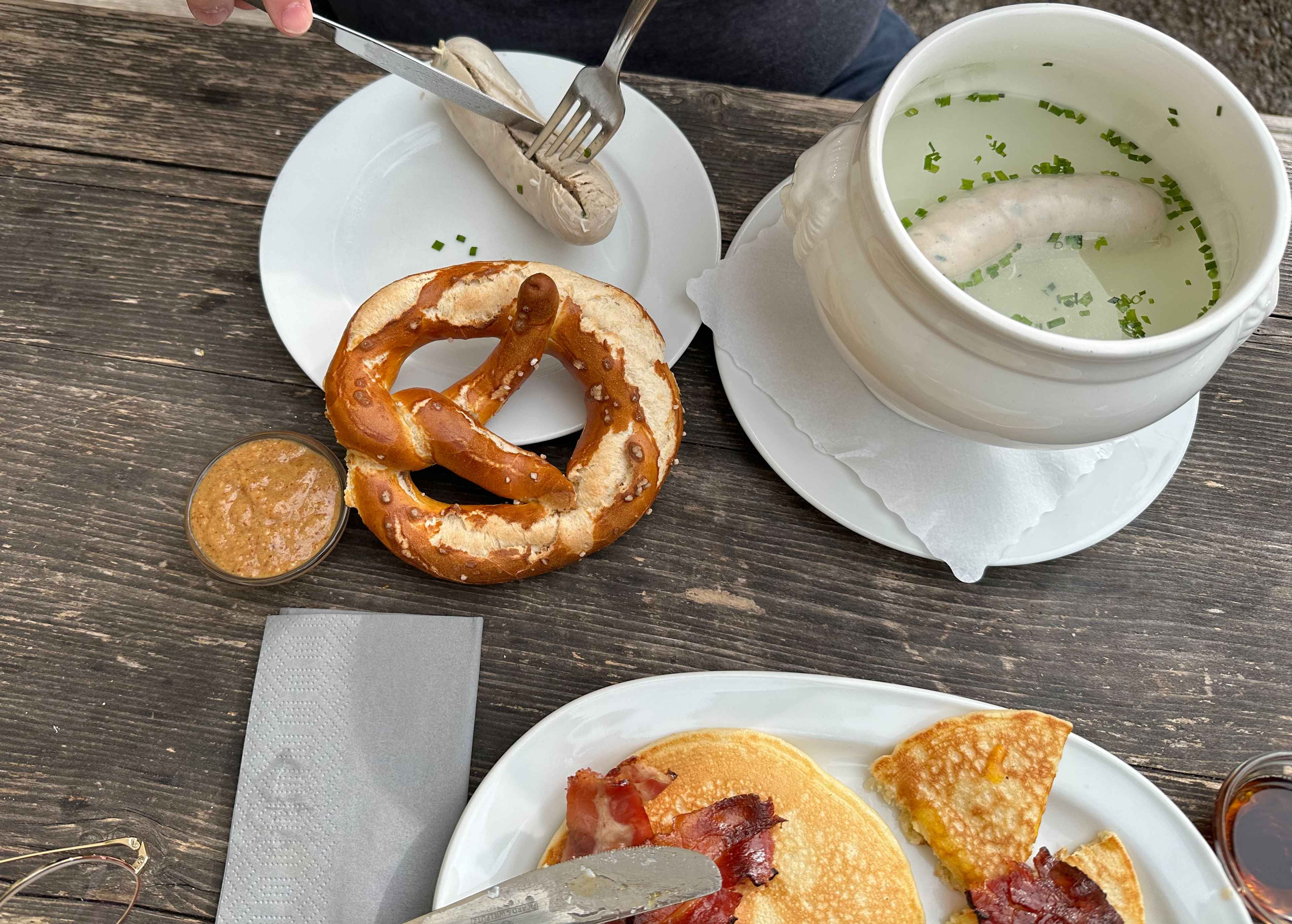 Blick von oben auf einen rustikalen Holztisch. Darauf Teller mit Weißwurst, Brezel und Pancakes.