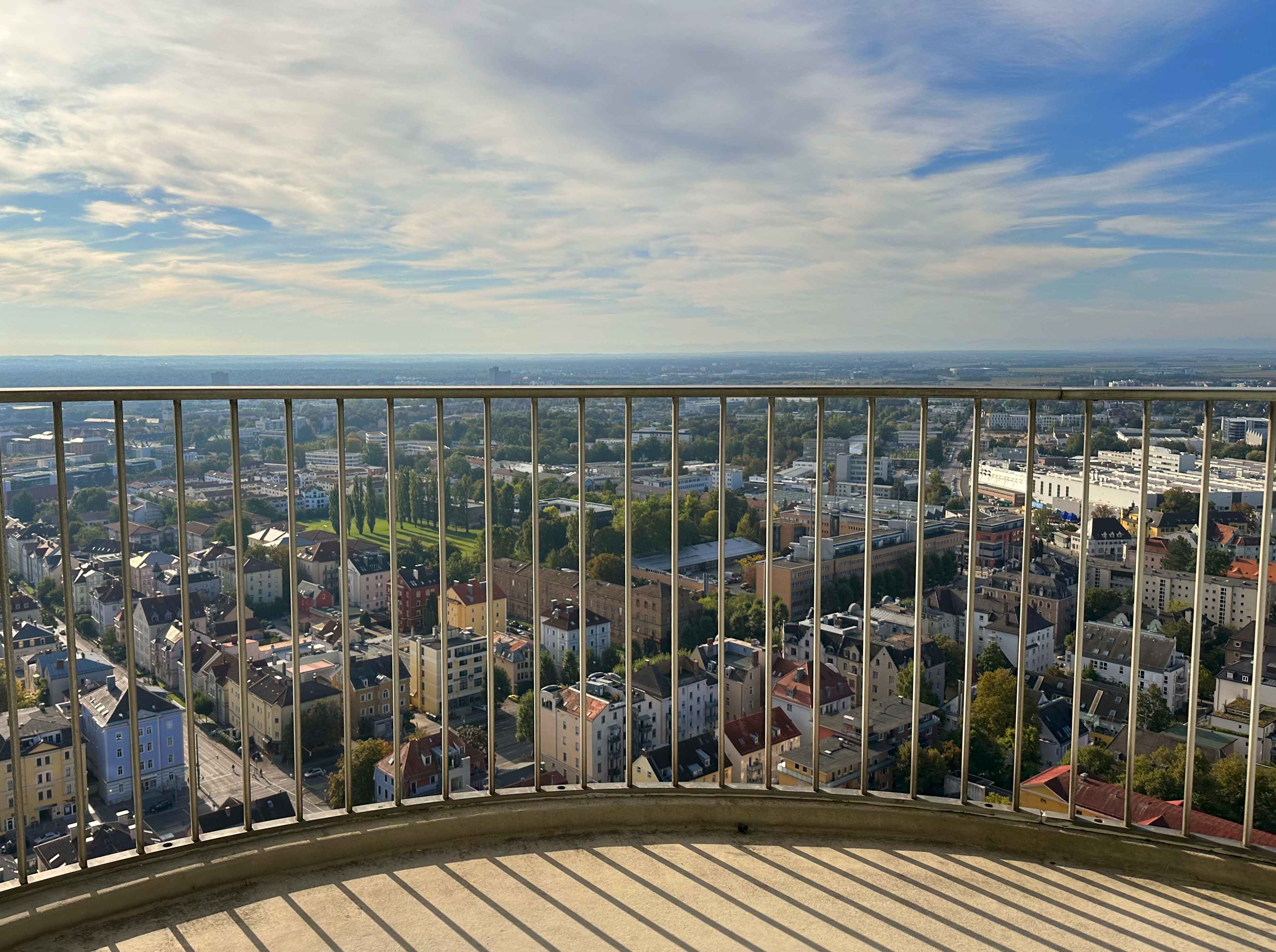 Blick durch das Mettallgelände des Balkons im 32. Stock auf die Augsburger Stadtlandschaft mit Gebäuden und Parkanlagen.