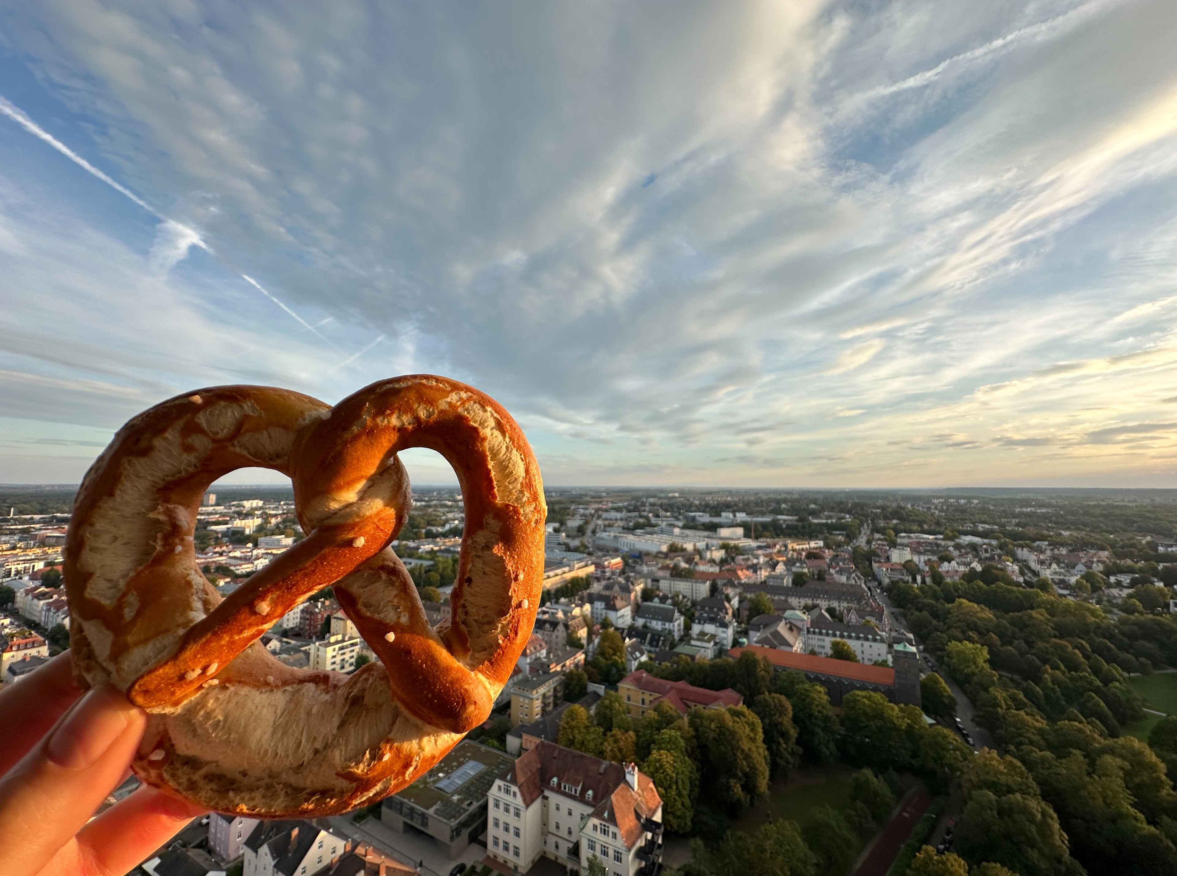 Eine Hand hält eine Brezel, dahinter ein Panorama der Stadt Augsburg von oben, mit sonnigem Himmel, Wolken und Flugstreifen.
