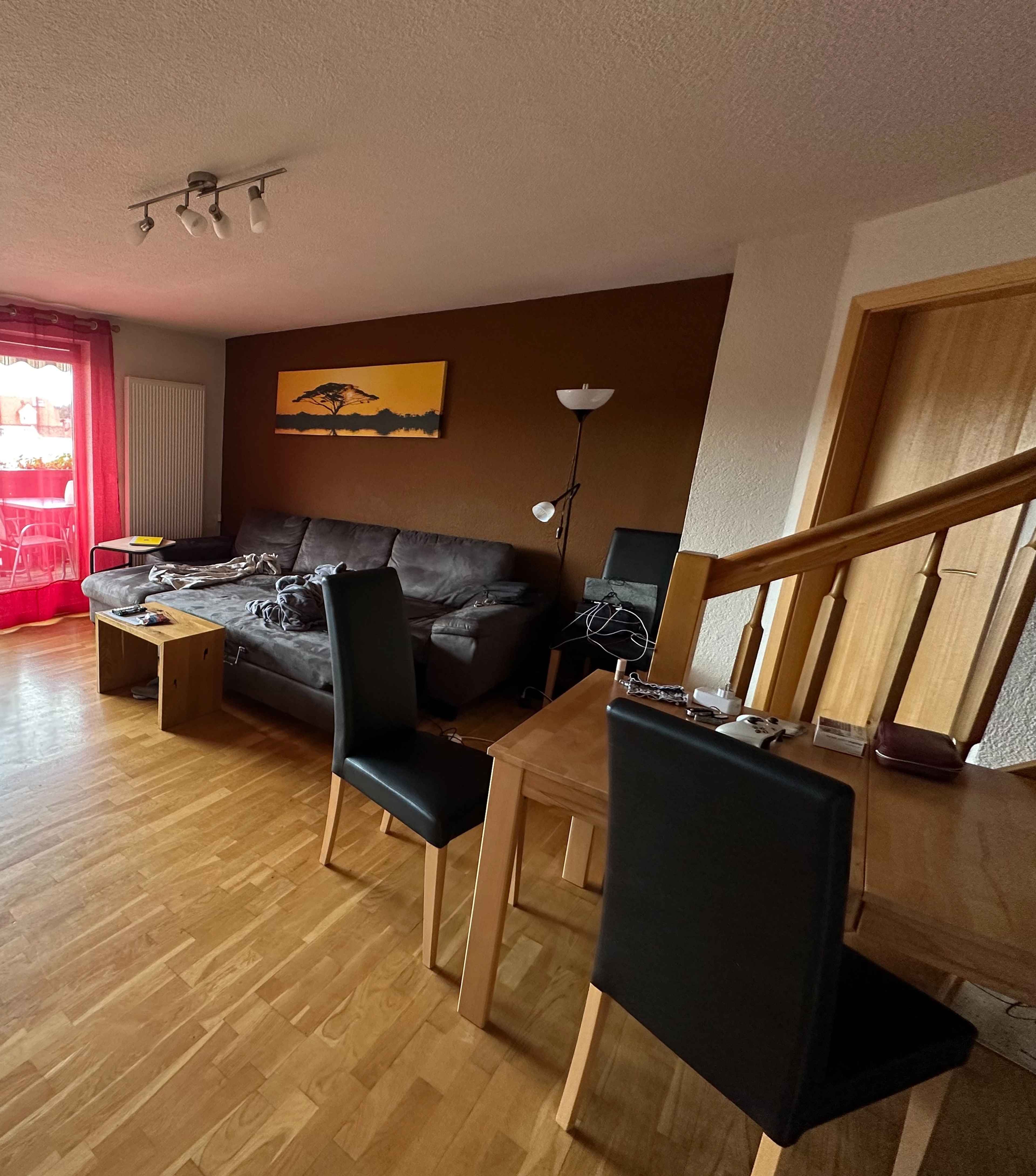 Ein Wohnzimmer mit brauner Wand, grauem Sofa, Esstisch und Stühlen. Im Hintergrund ein großes Fenster mit roten Vorhängen.