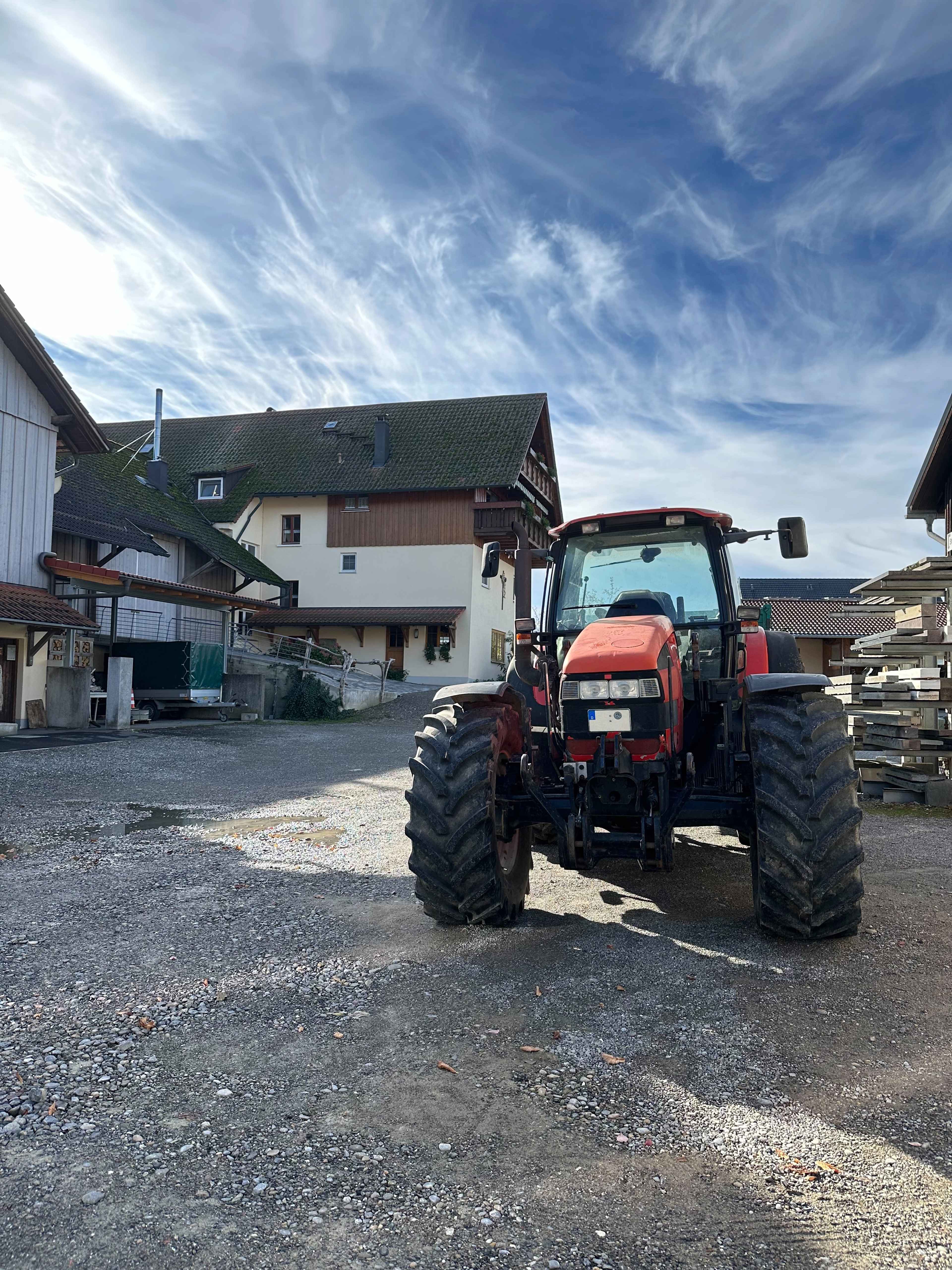 Frontalsicht auf einen roten Traktor auf einem Schotterplatz vor einem seitlich sichtbaren Bauernhaus unter blauem Himmel.