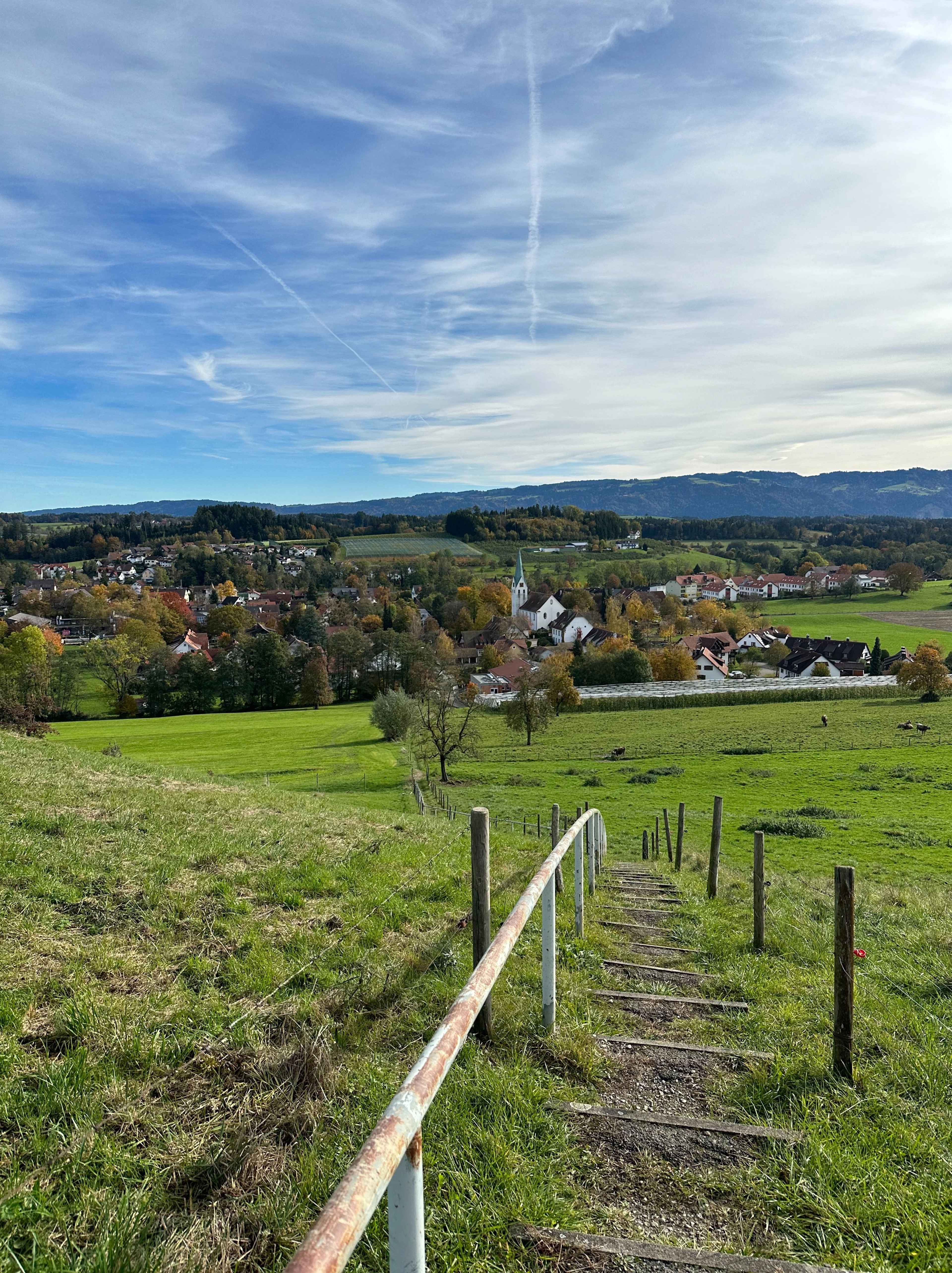 Sicht vom grünen Hügel auf ein Dorf unter blauem Himmer. In der Mitte führen Stufen mit Holzgeländer die Wiese hinunter.