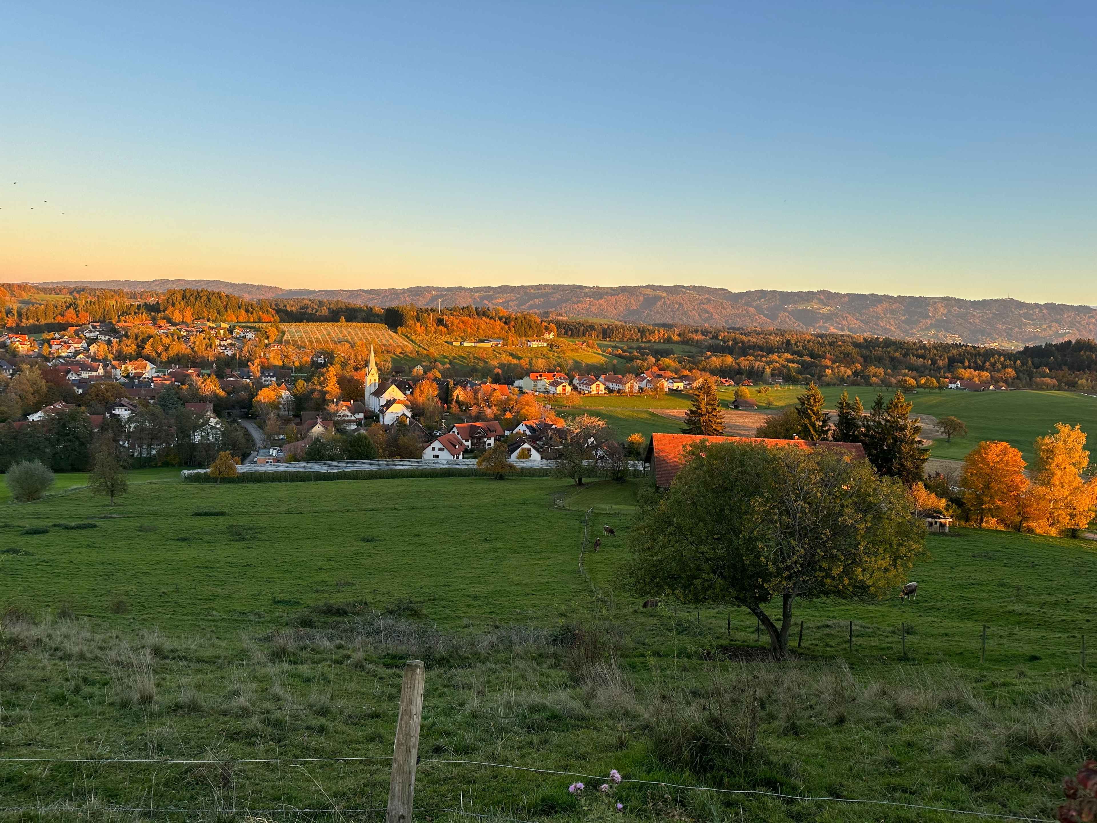 Herbstliche Landschaft im Abendrot mit Blick auf ein Dorf umgeben von grünen Wiesen und Bäumen in herbstlichen Farben.