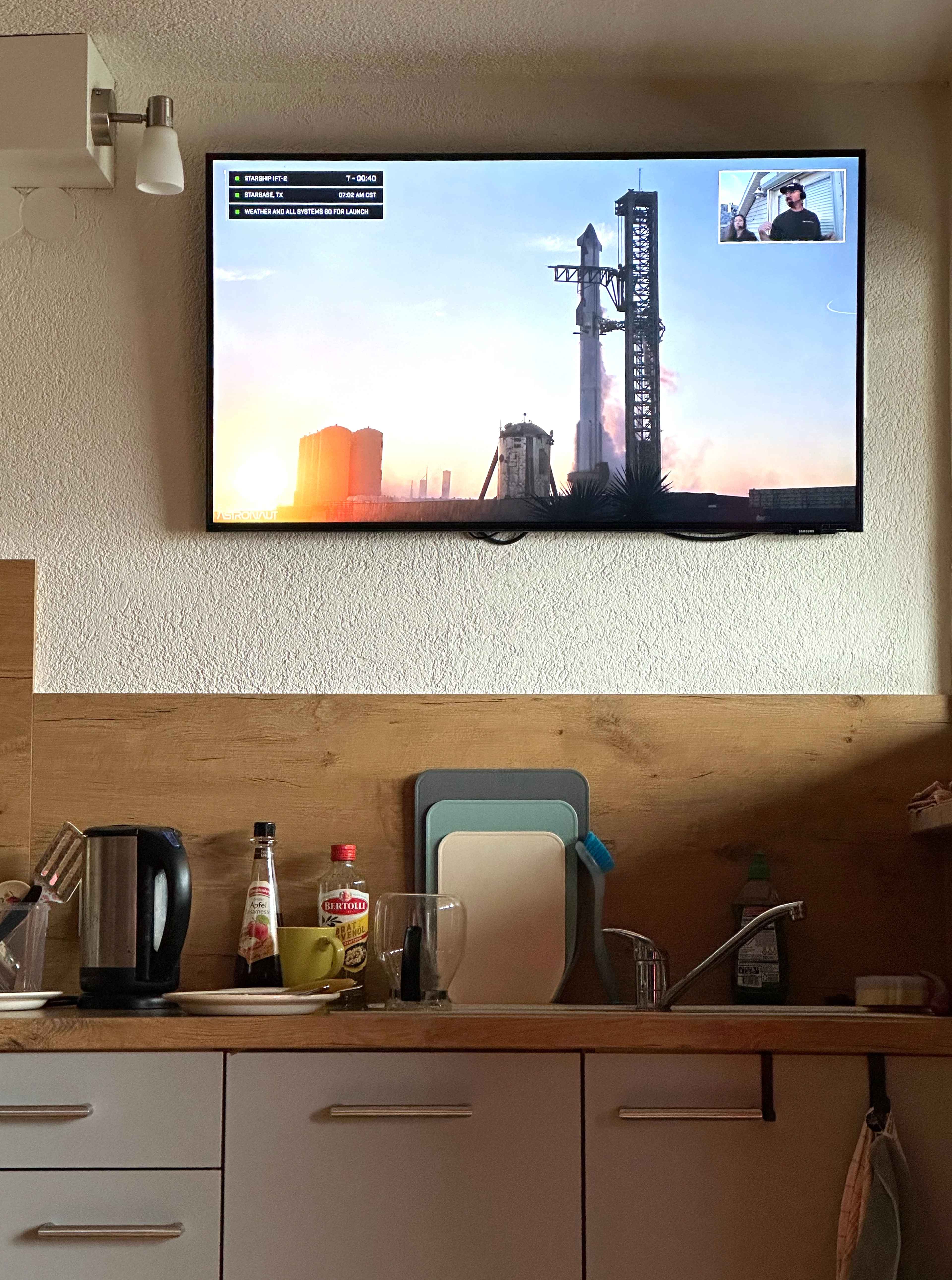 Ein Fernseher zeigt einen Raketenstart mit Countdown. Darunter eine Küchentheke mit Wasserkocher und Schneidebrettern.