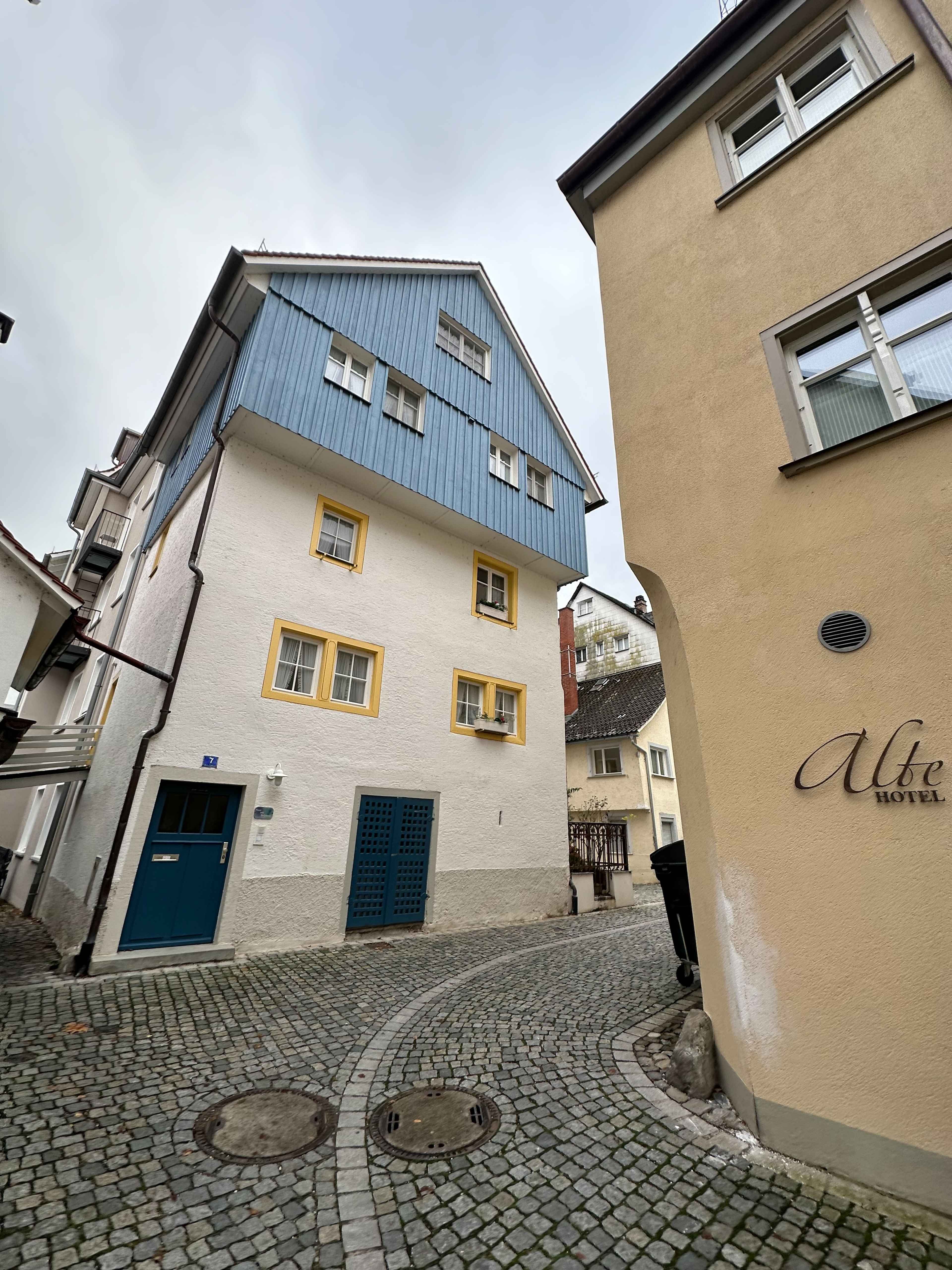 Eine schmale Gasse mit Kopfsteinpflaster, flankiert von Häusern. Ein Haus hat eine blaue Holzfassade und gelbe Fensterrahmen.