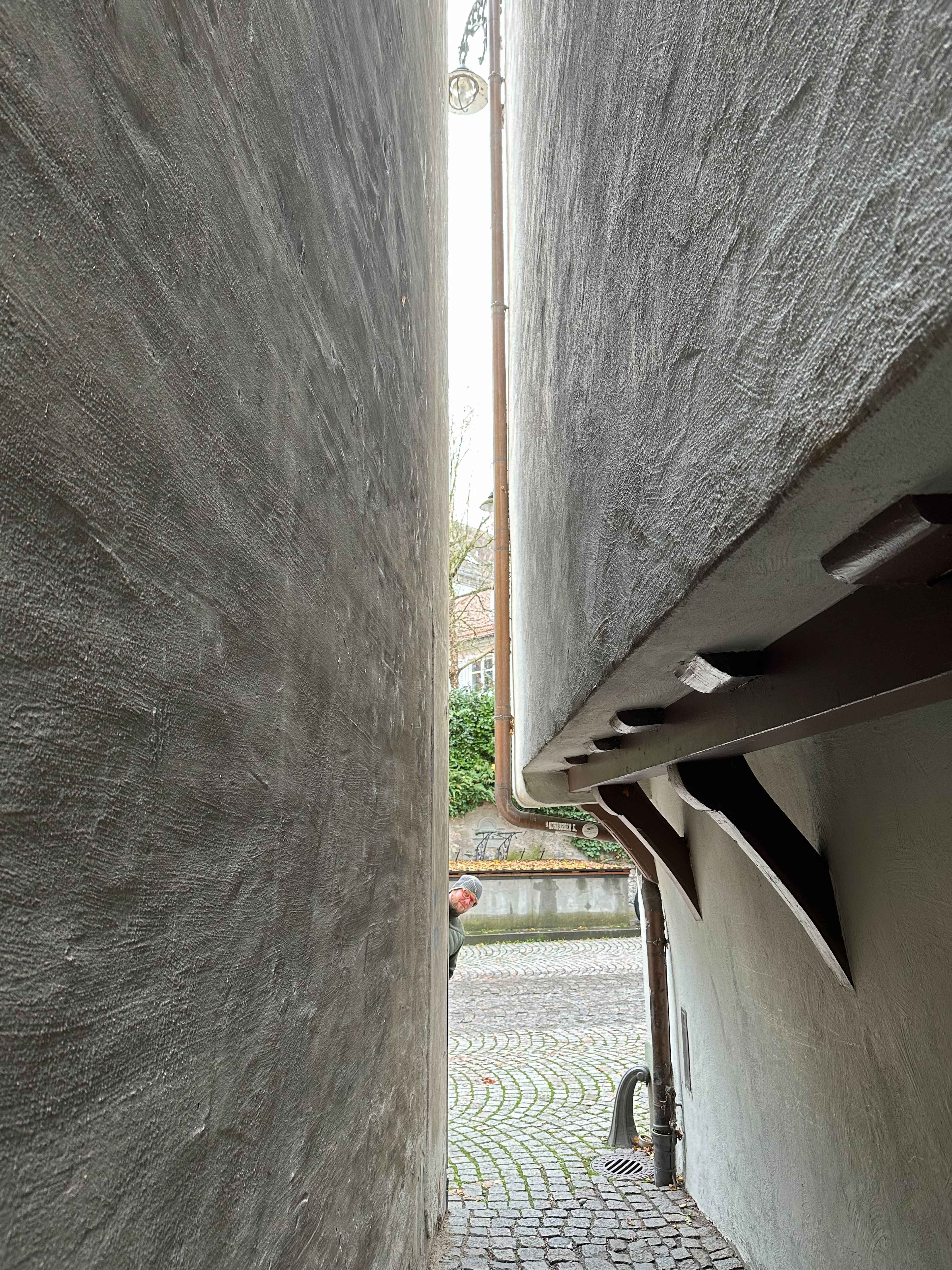Ein enger Spalt zwischen zwei grauen Hauswänden. Unten schaut ein Mann mit Mütze hervor, im Hintergrund Kopfsteinpflaster.