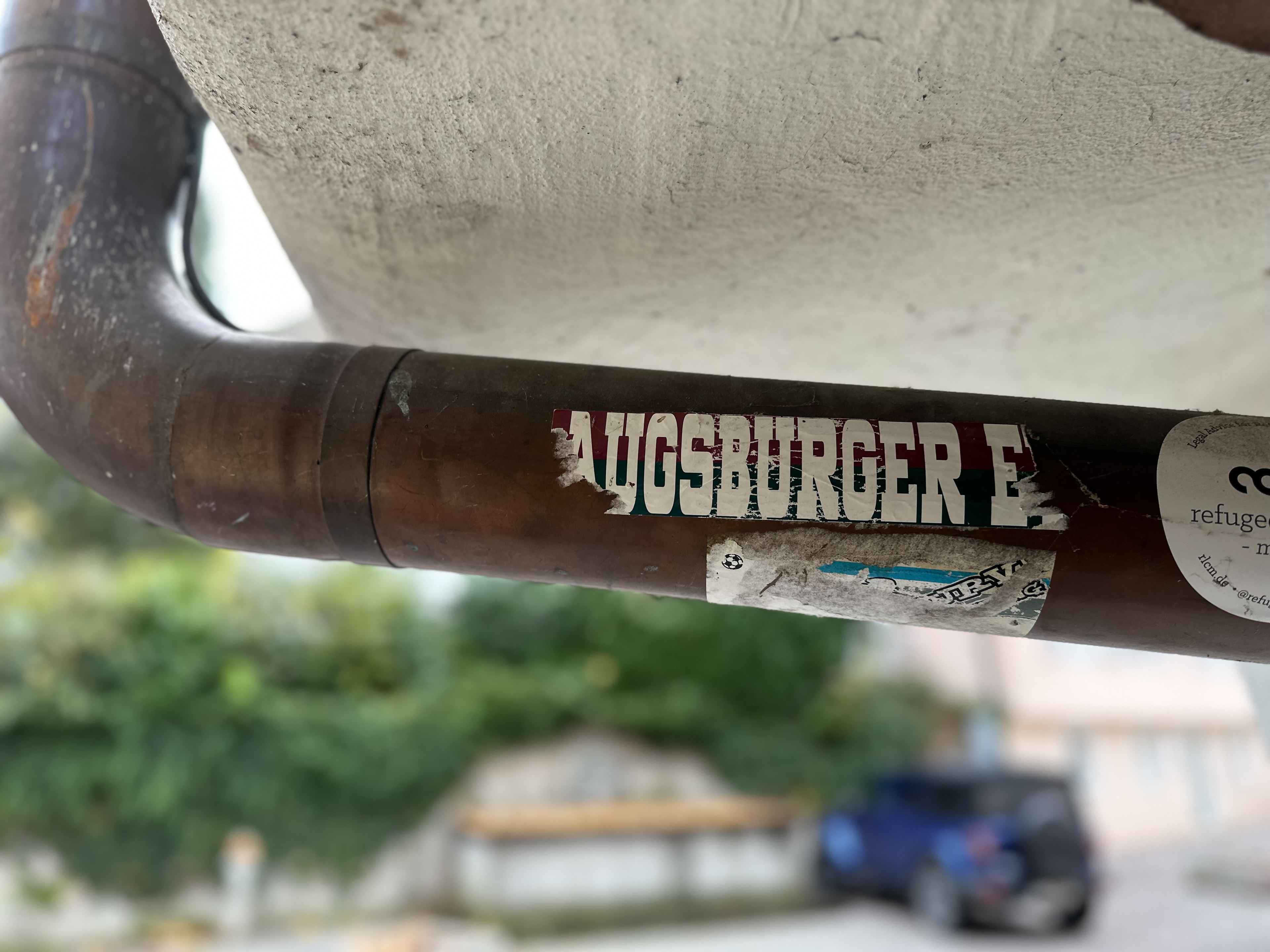 Ein verrostetes Metallrohr mit einem abgenutzten Aufkleber, auf dem "Augsburger E" steht. Unscharfer grüner Hintergrund.
