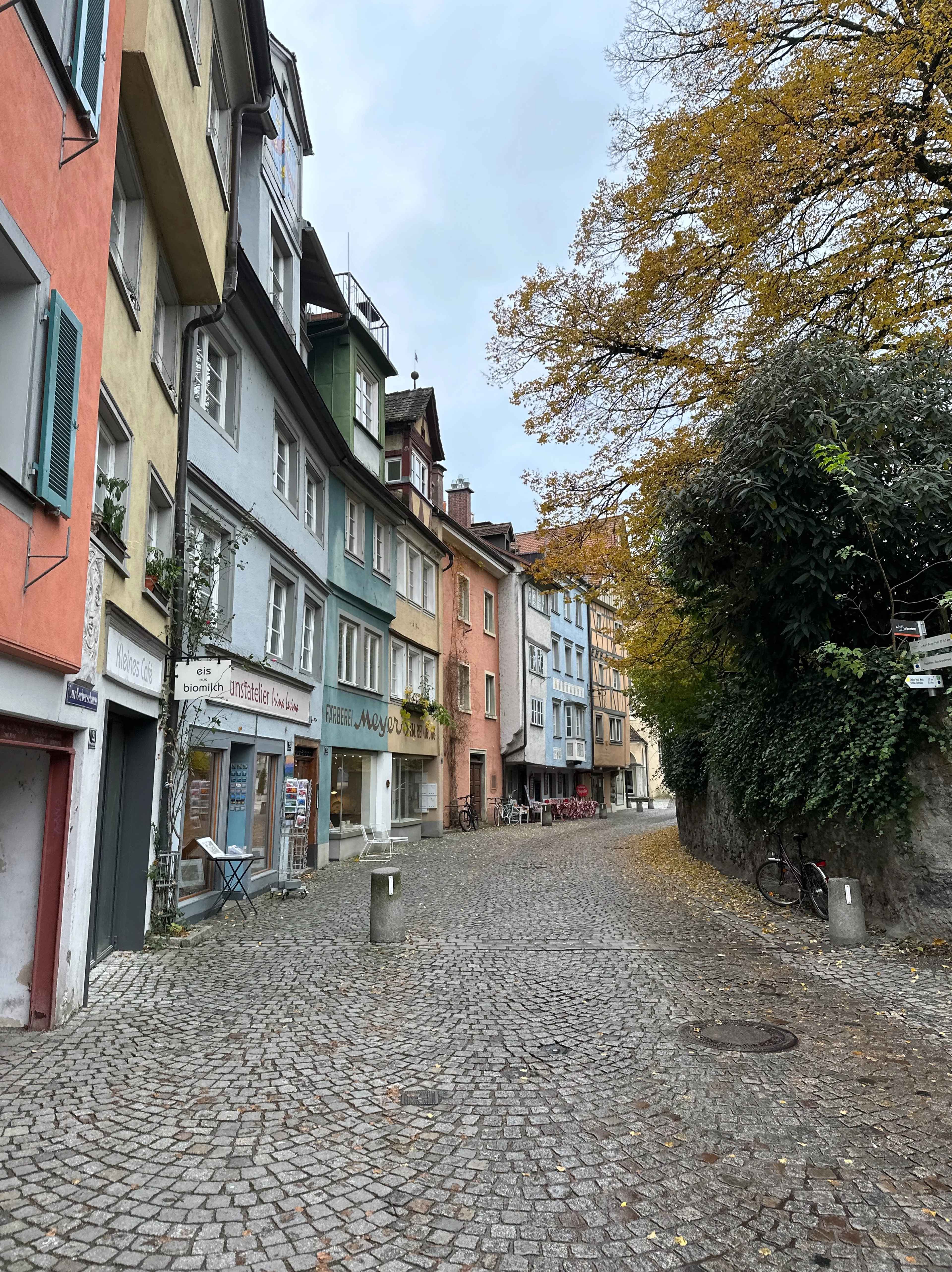 Eine gepflasterte Gasse in der Lindauer Altstadt, gesäumt von bunten historischen Häusern. Rechts Bäume mit Herbstlaub.