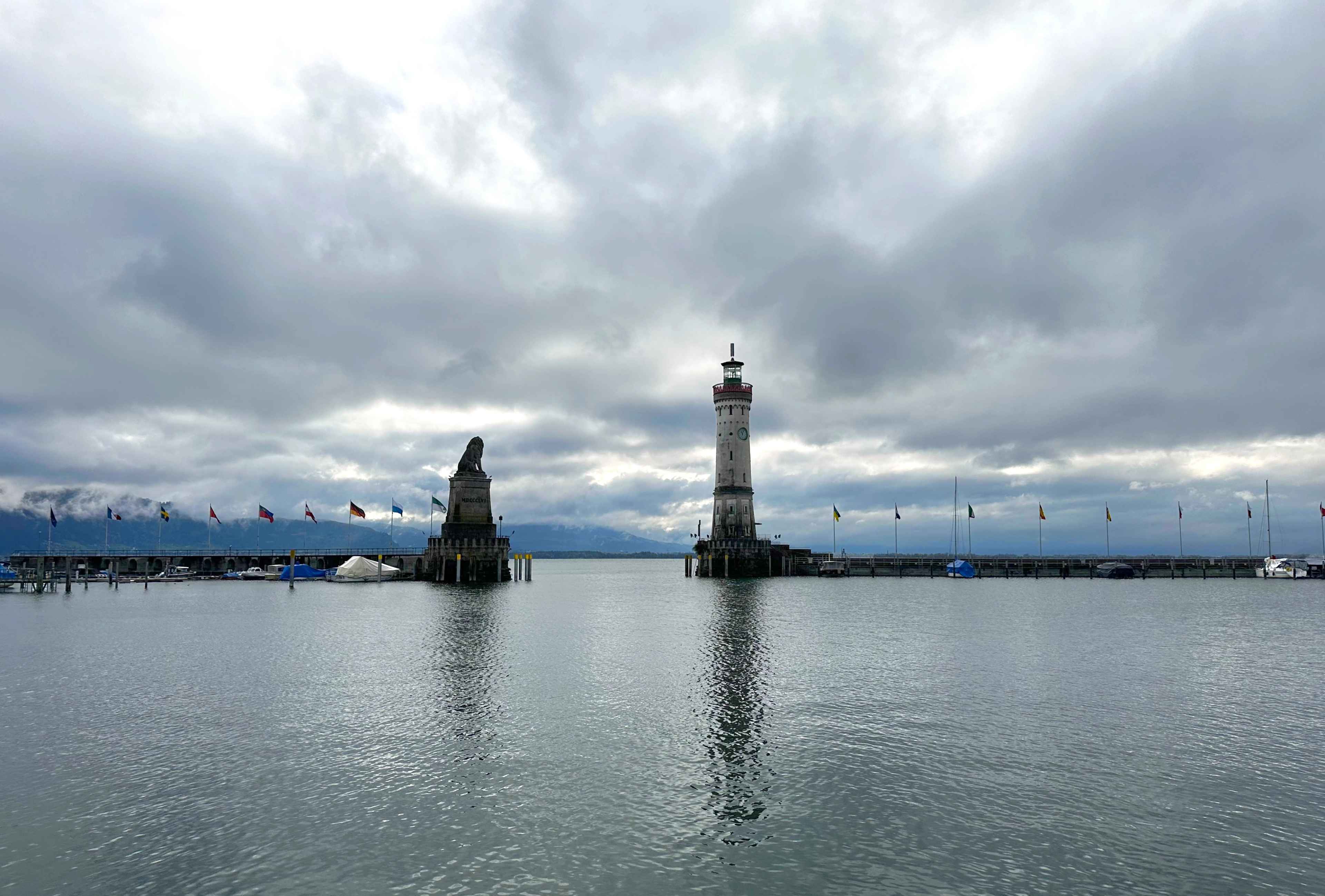 Die Hafeneinfahrt mit dem Lindauer Löwen und Leuchtturm. Dazwischen Flaggen, dahinter der wolkenverhangene Bodensee.