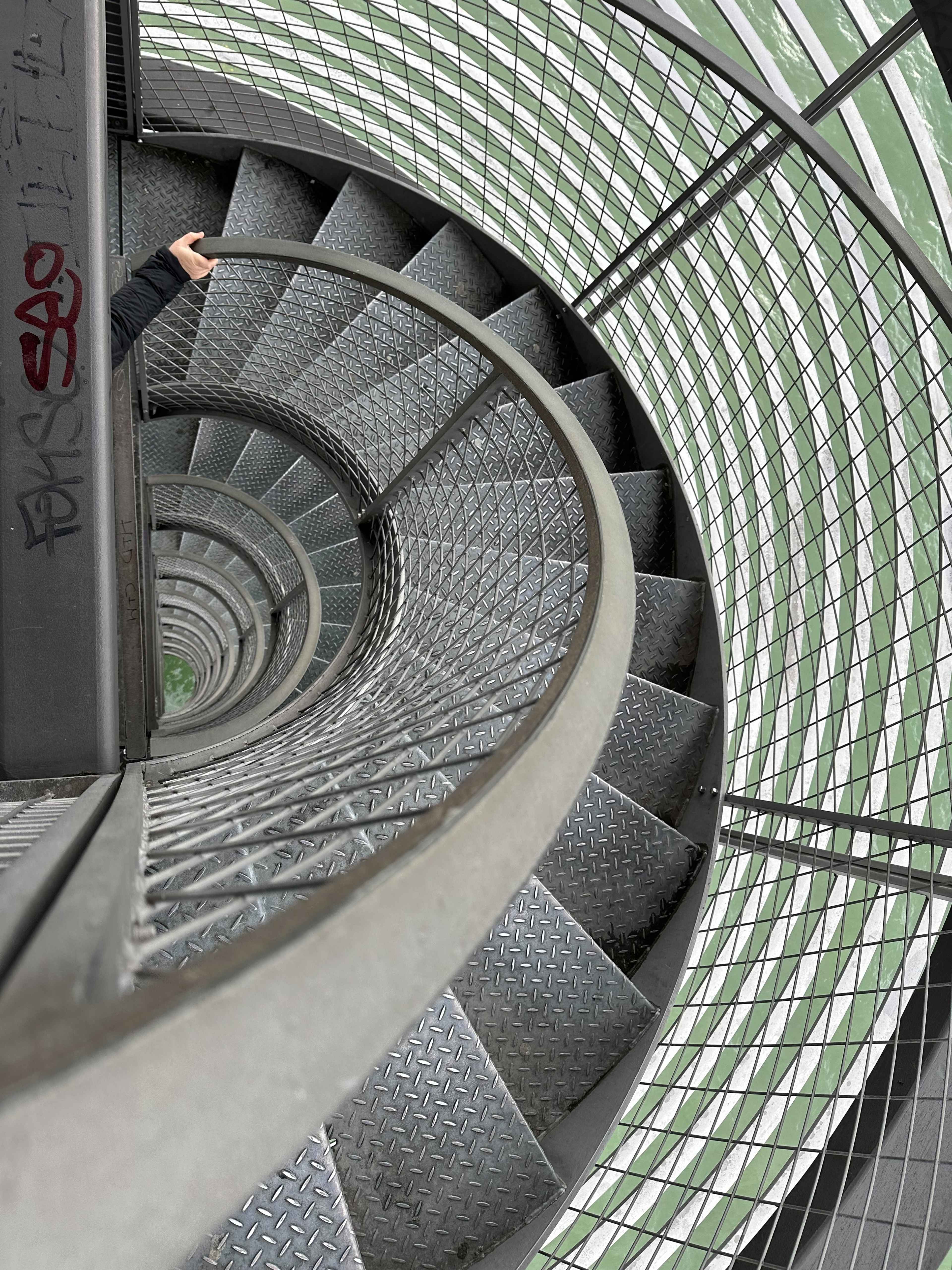 Eine spiralförmige Metalltreppe mit Gittergeländer windet sich nach unten. Eine Hand hält sich am Geländer fest.