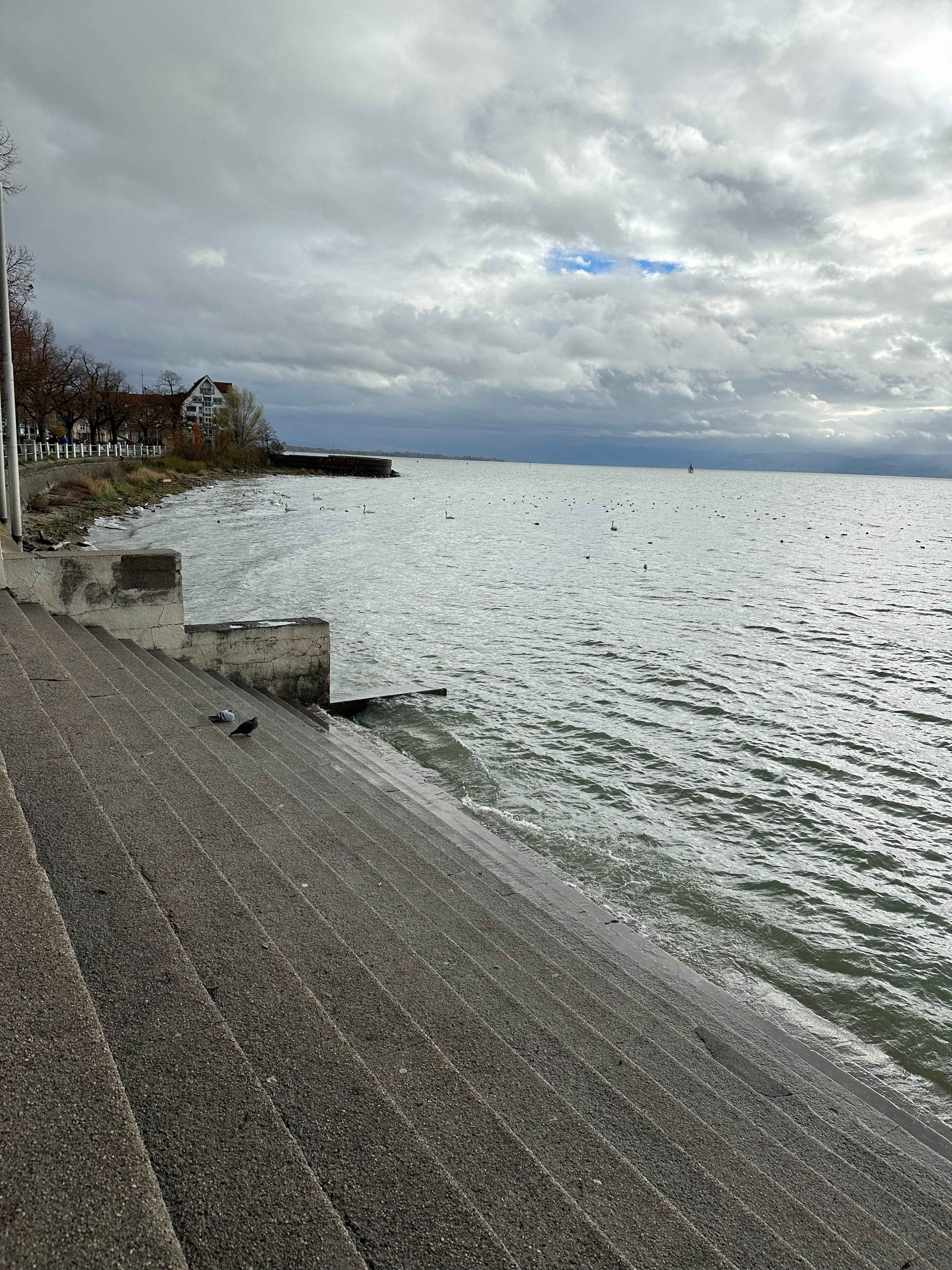 Stufen führen zum Ufer eines Sees, der sich weit bis zum Horizont erstreckt, unter einem wolkigen Himmel.