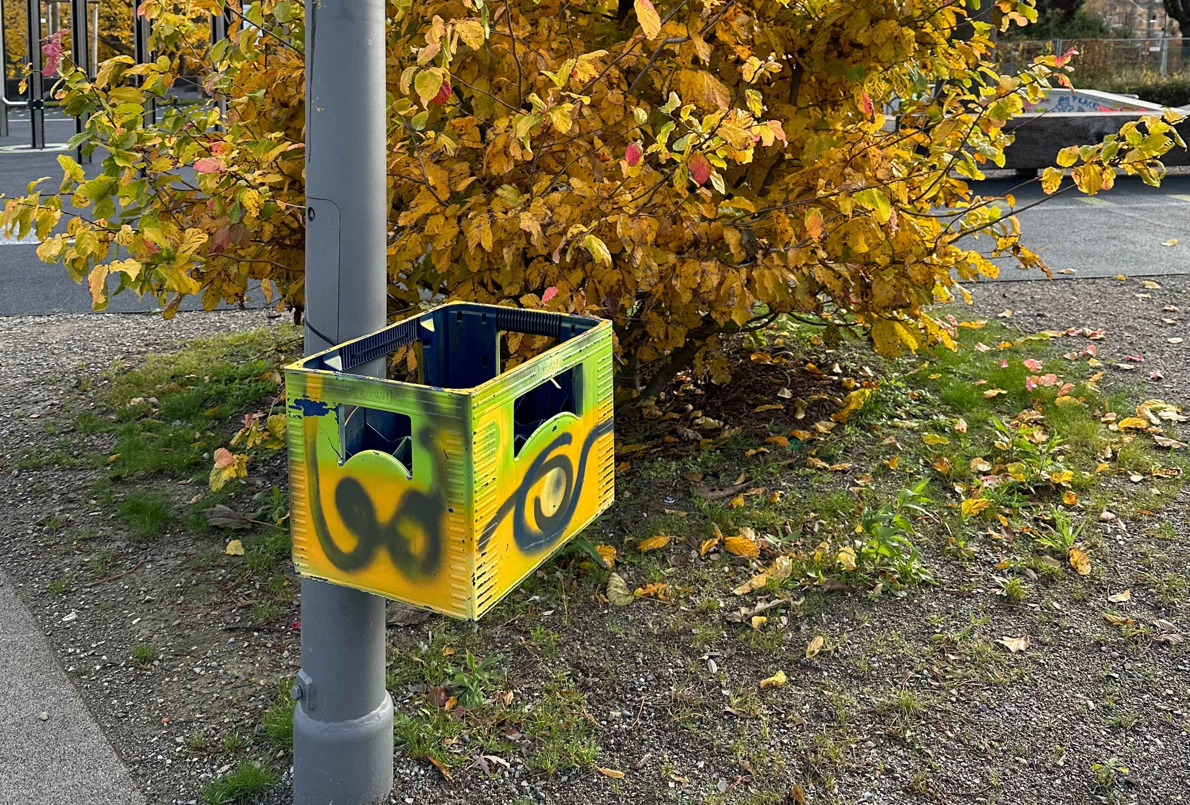 Ein gelb-grüner, bunt bemalter Bierkasten angebunden an eine Laterne, dahinter ein Busch mit gelbem Herbstlaub.