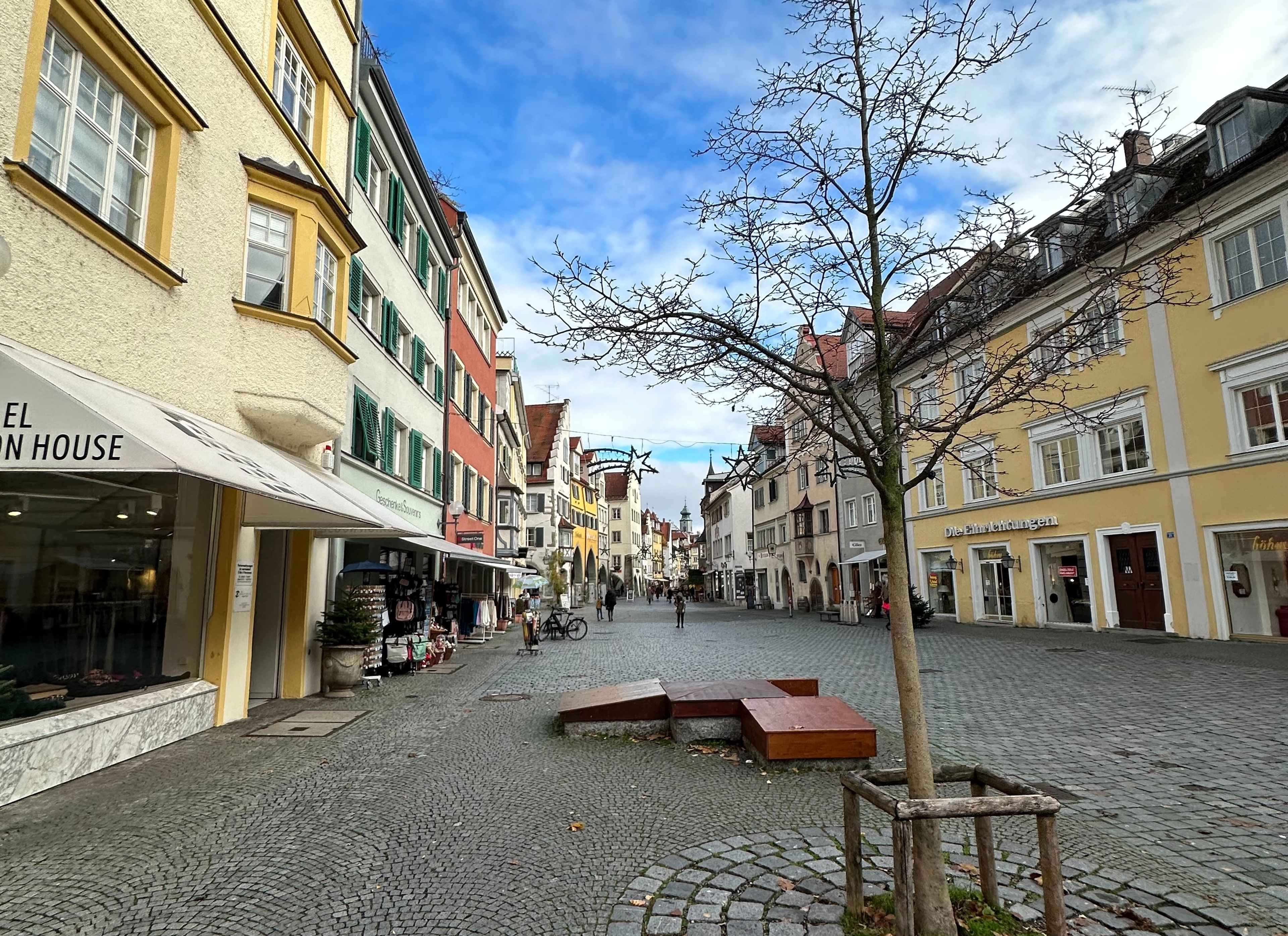 Eine Kopfsteinpflasterstraße mit bunten Häusern und kleinen Geschäften. Ein Baum steht in der Mitte eines offenen Platzes.