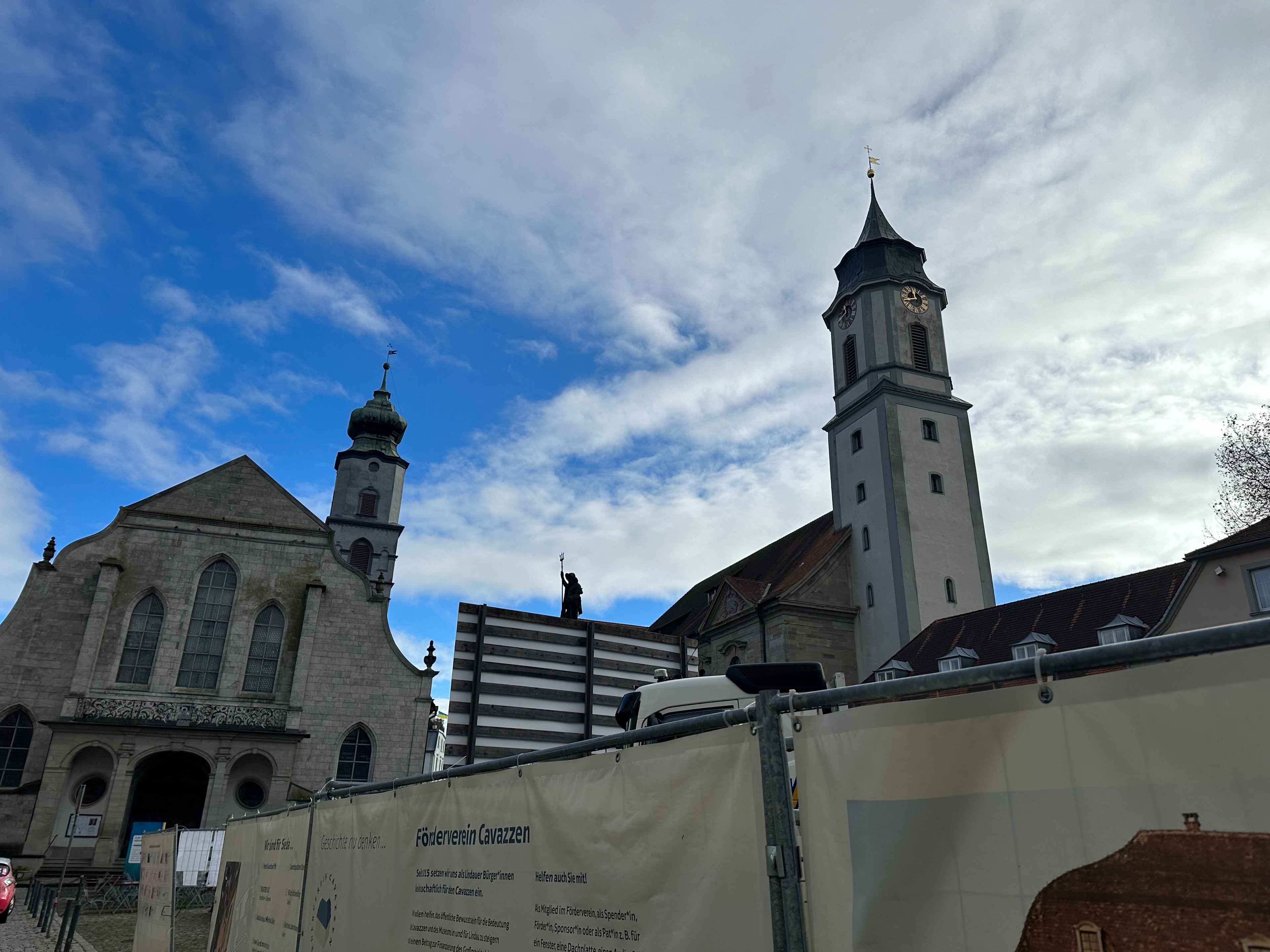 Kirchengebäude mit zwei Türmen und Bauzaun im Vordergrund, blauer Himmel mit weißen Wolken im Hintergrund.