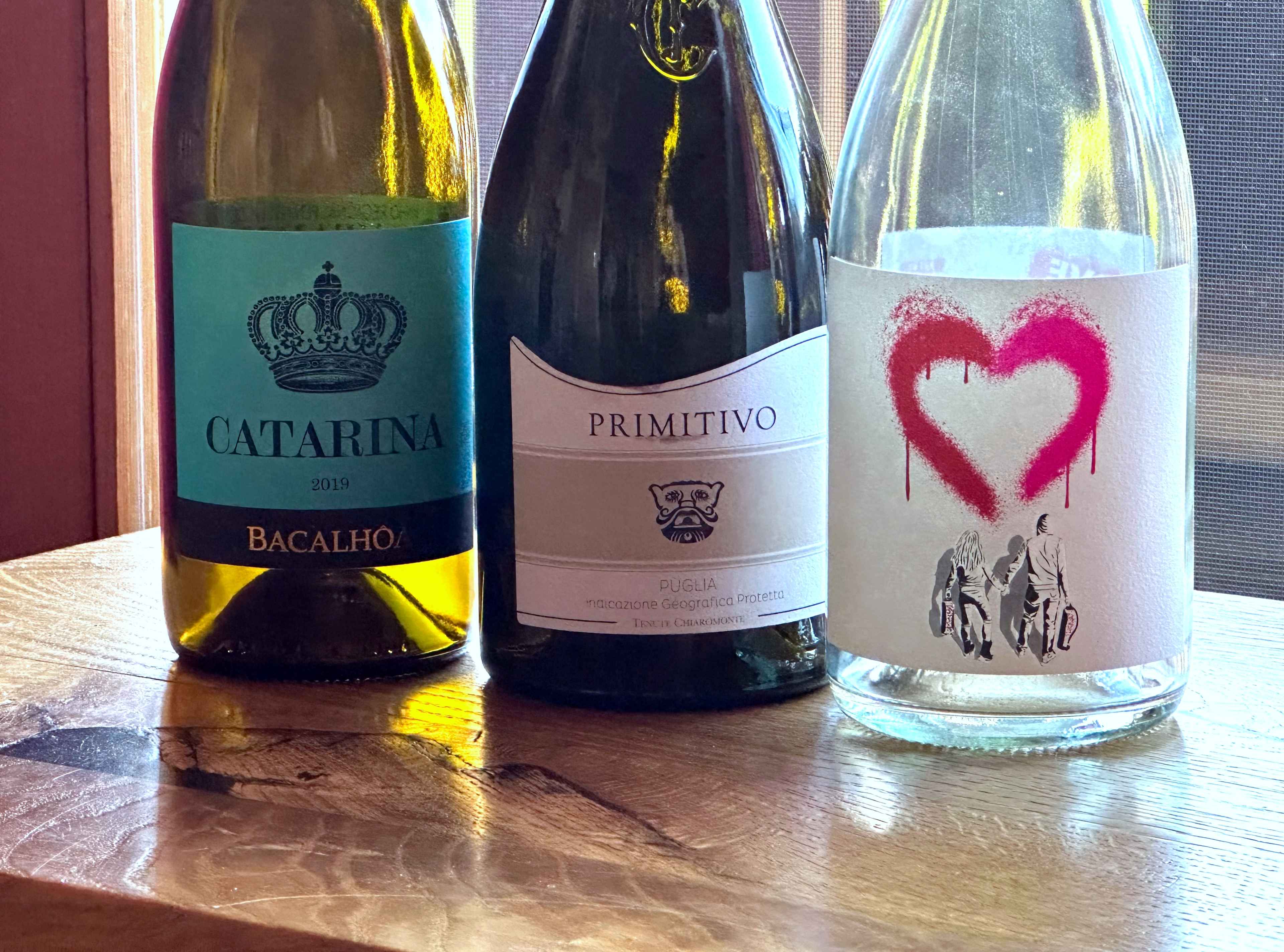 Drei Weinflaschen auf einem Holztisch: "Catarina", "Primitivo" und eine Flasche mit einem roten Herzmotiv auf dem Etikett.
