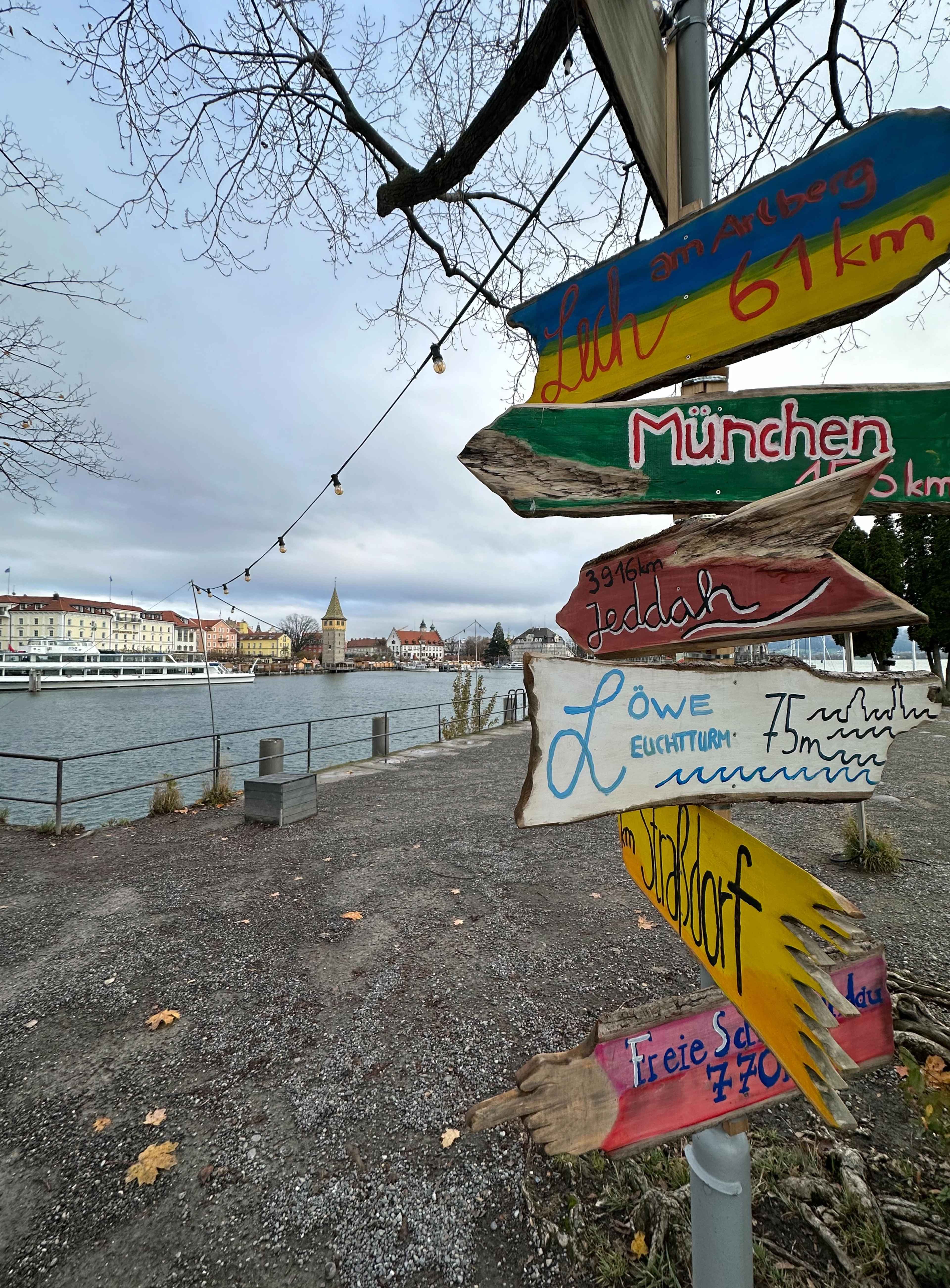 Ein bunter Wegweiser mit handbeschrifteten Entfernungen zu Orten wie München und Jeddah, im Hintergrund eine Hafenpromenade.