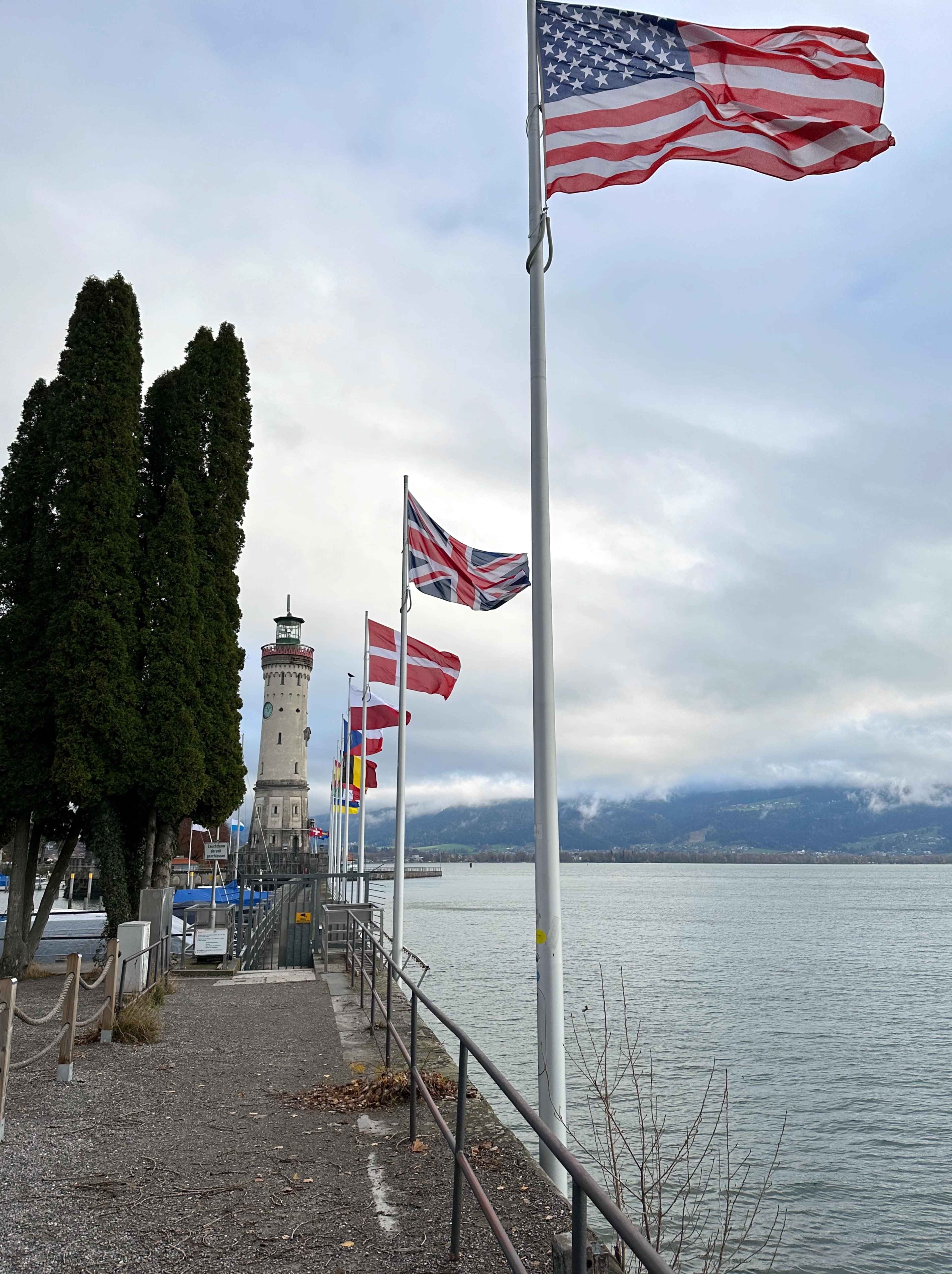 Der Lindauer Leuchtturm am Hafen, umgeben von Flaggen verschiedener Länder, mit See und Bergen im Hintergrund.
