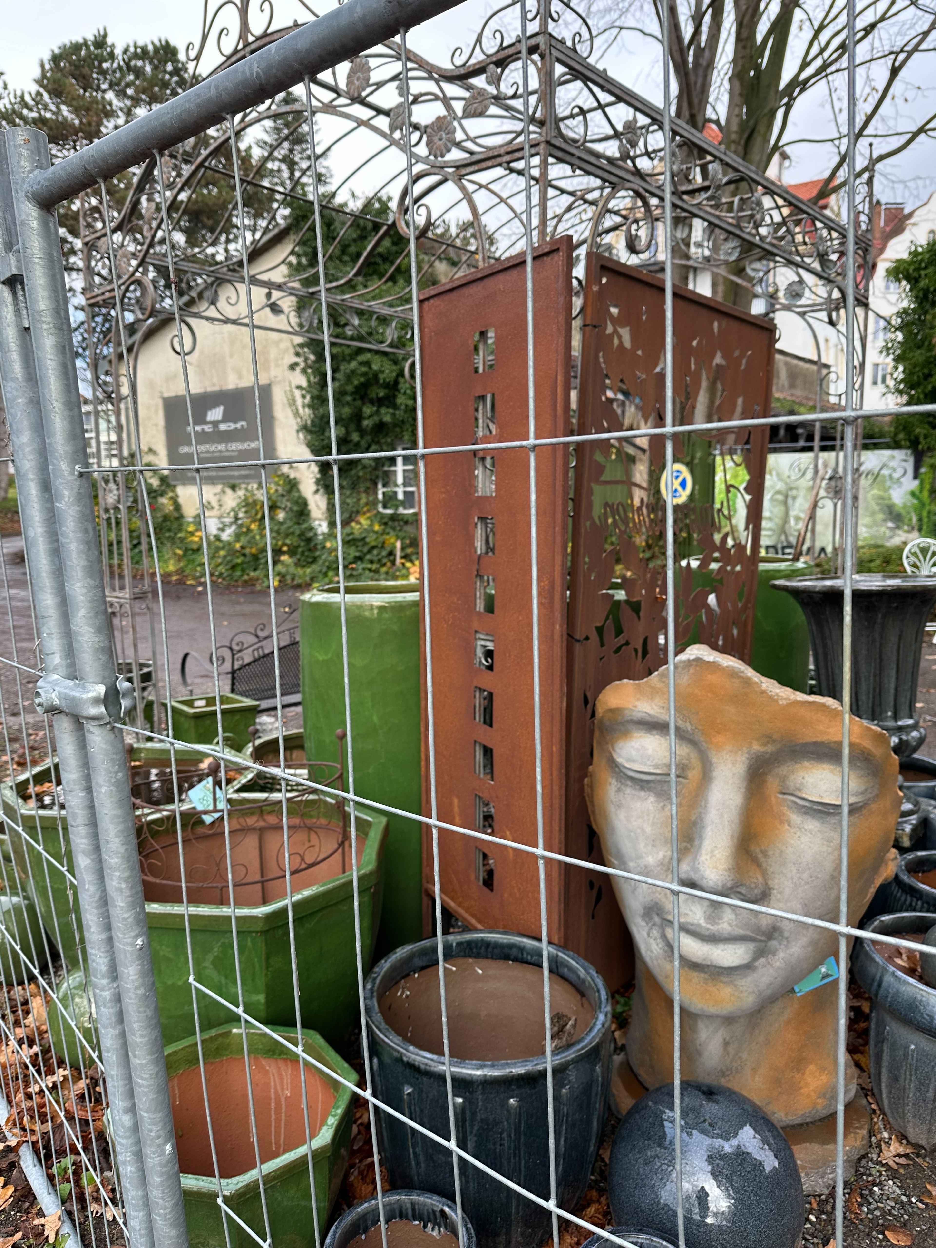 Eine Gartenfigur mit geschlossenen Augen, umgeben von Töpfen und Dekorationen, hinter einem Gitterzaun.