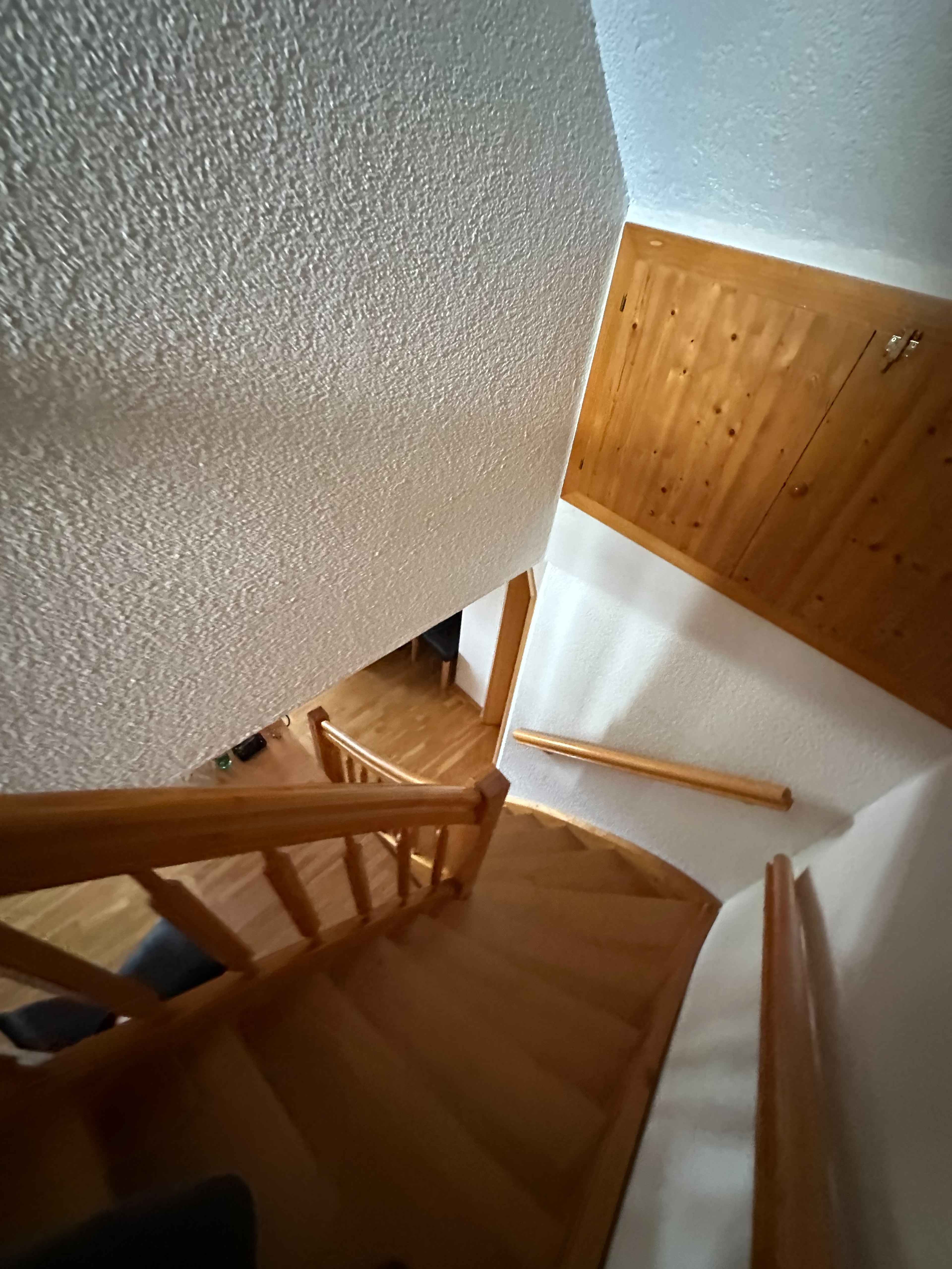 Eine Holztreppe mit Geländer führt nach unten in einen Raum mit Holzboden, helle Wände und eine Decke aus Holz und Putz.
