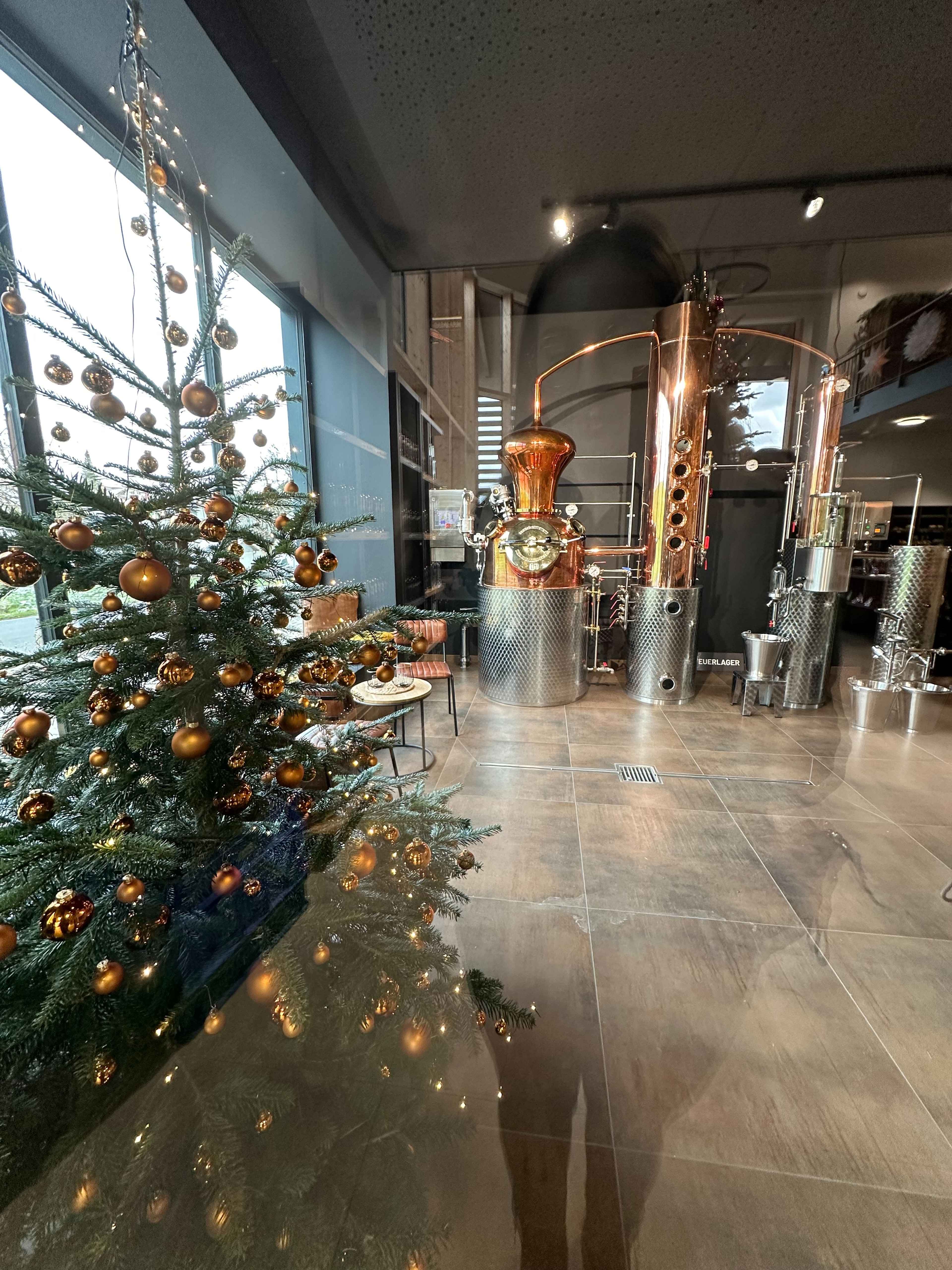 Blick durch eine Scheibe auf kupferne Destillationsgeräte einer Brennerei, daneben ein Weihnachtsbaum mit goldenen Kugeln.