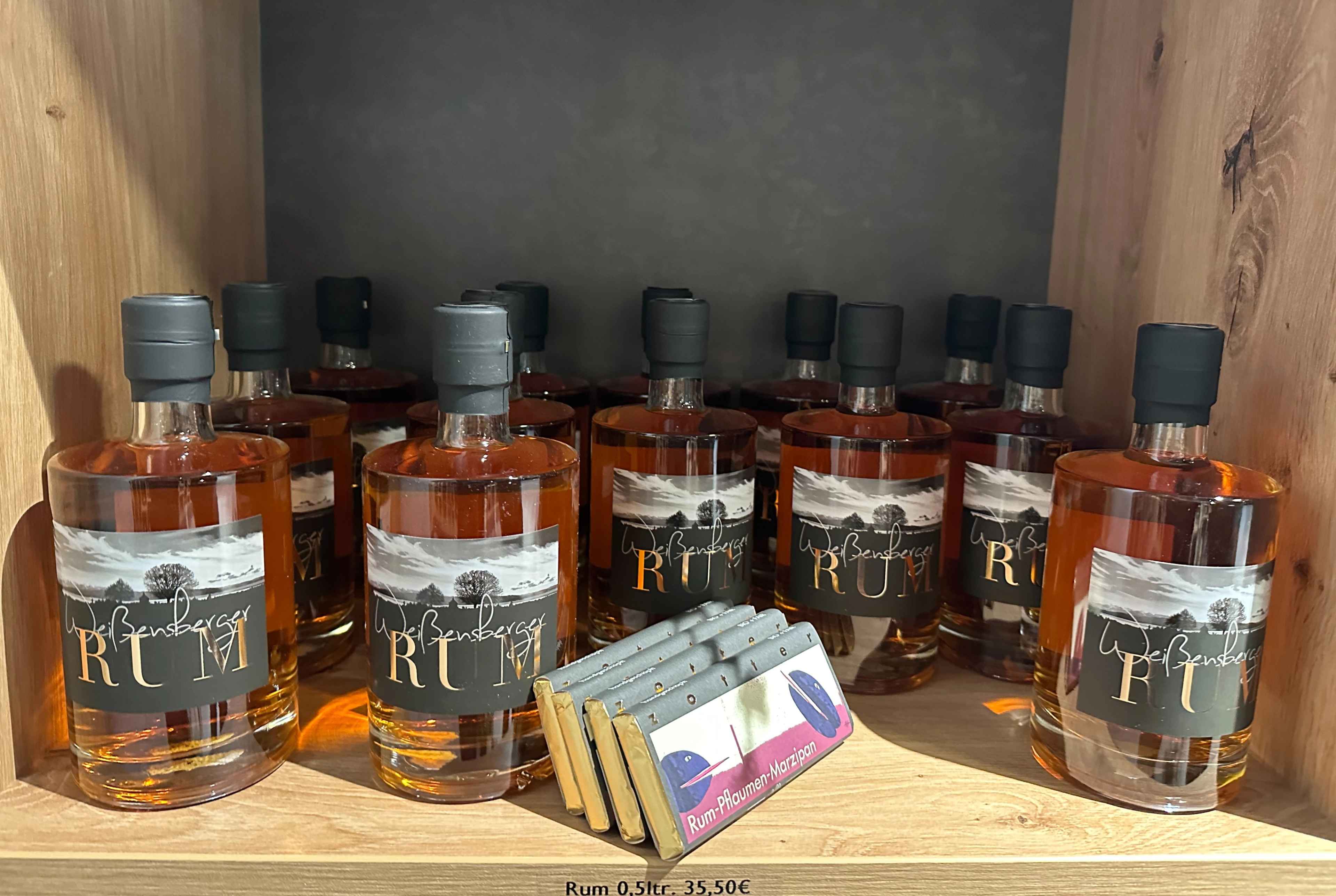 Regal mit mehreren Flaschen regionalem Rum, beschriftet mit 'Weißenberger Rum', daneben kleine Schokoladentafeln.