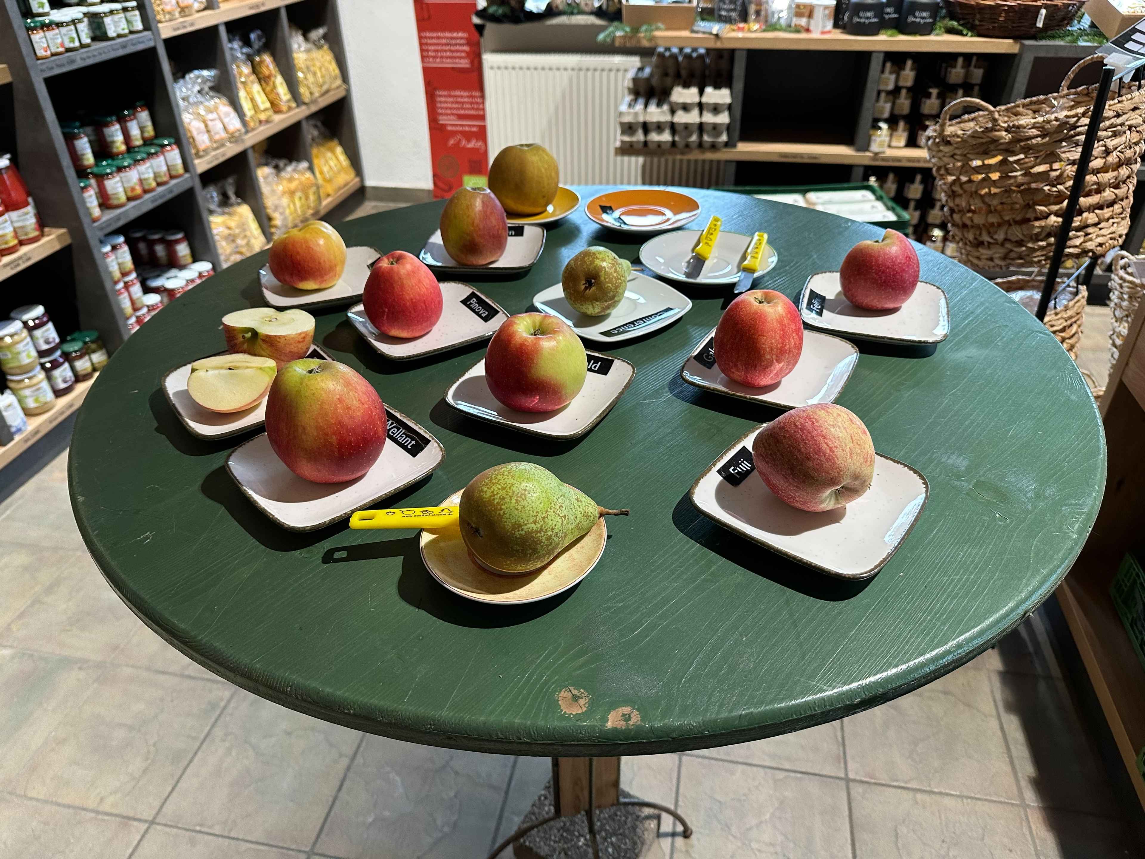 Tisch mit verschiedenen Apfel- und Birnensorten auf Tellern auf einem grünen Tisch, jede Sorte mit einem Schild beschriftet.